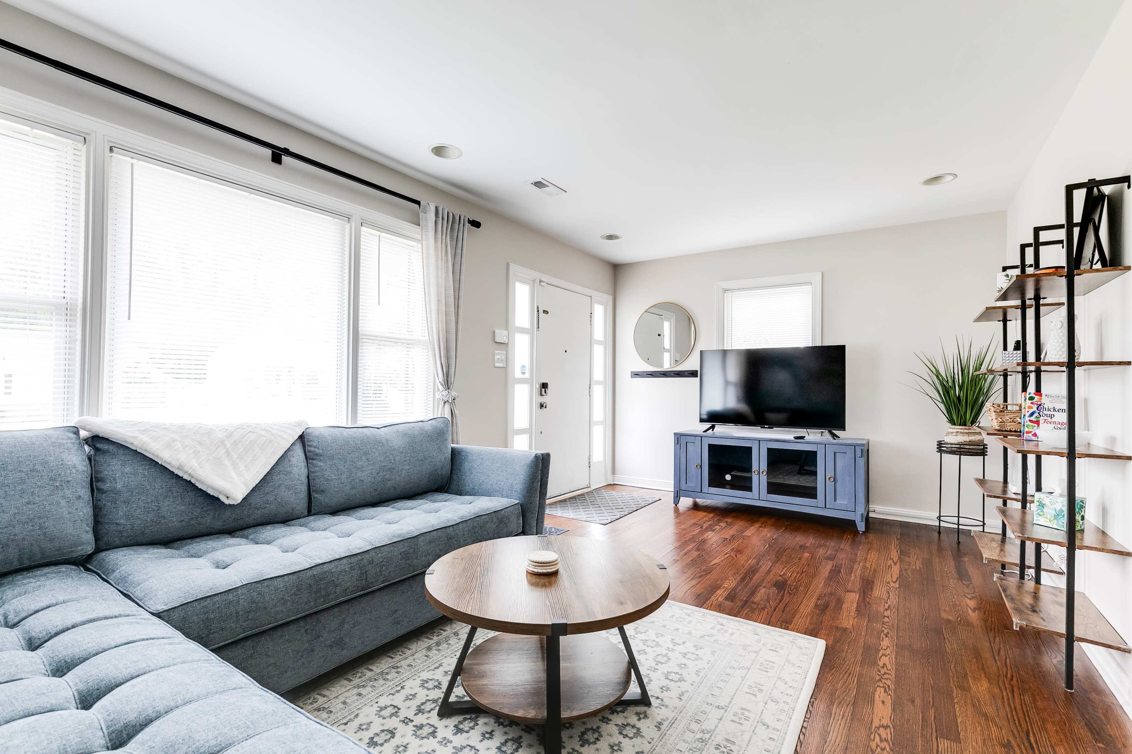 The living room features a blue sectional sofa, a round wooden coffee table, and a television on a console, all set against a backdrop of large windows and light-colored walls.
