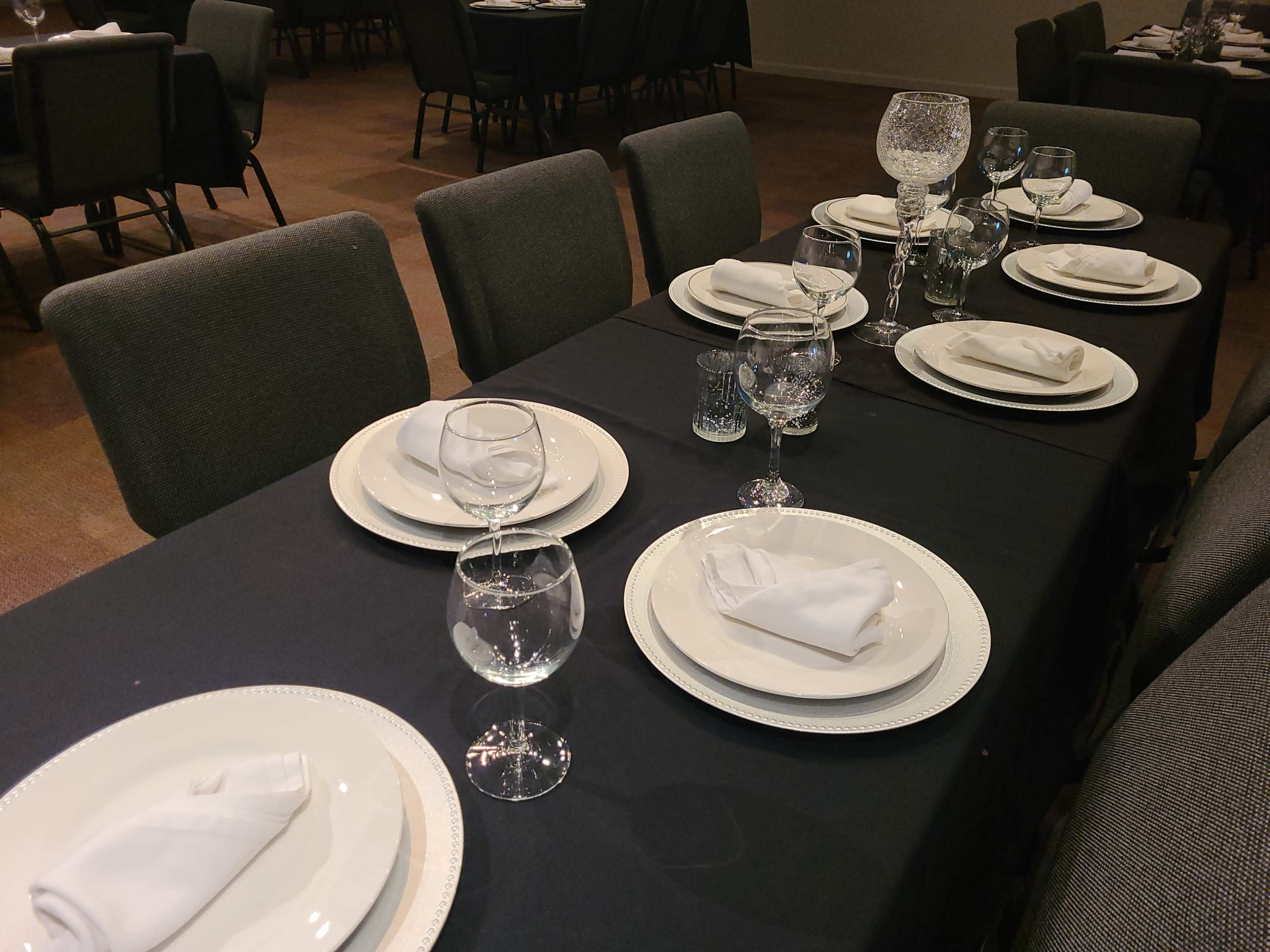 A long dining table is set with white plates, folded napkins, and various types of glassware on a black tablecloth.