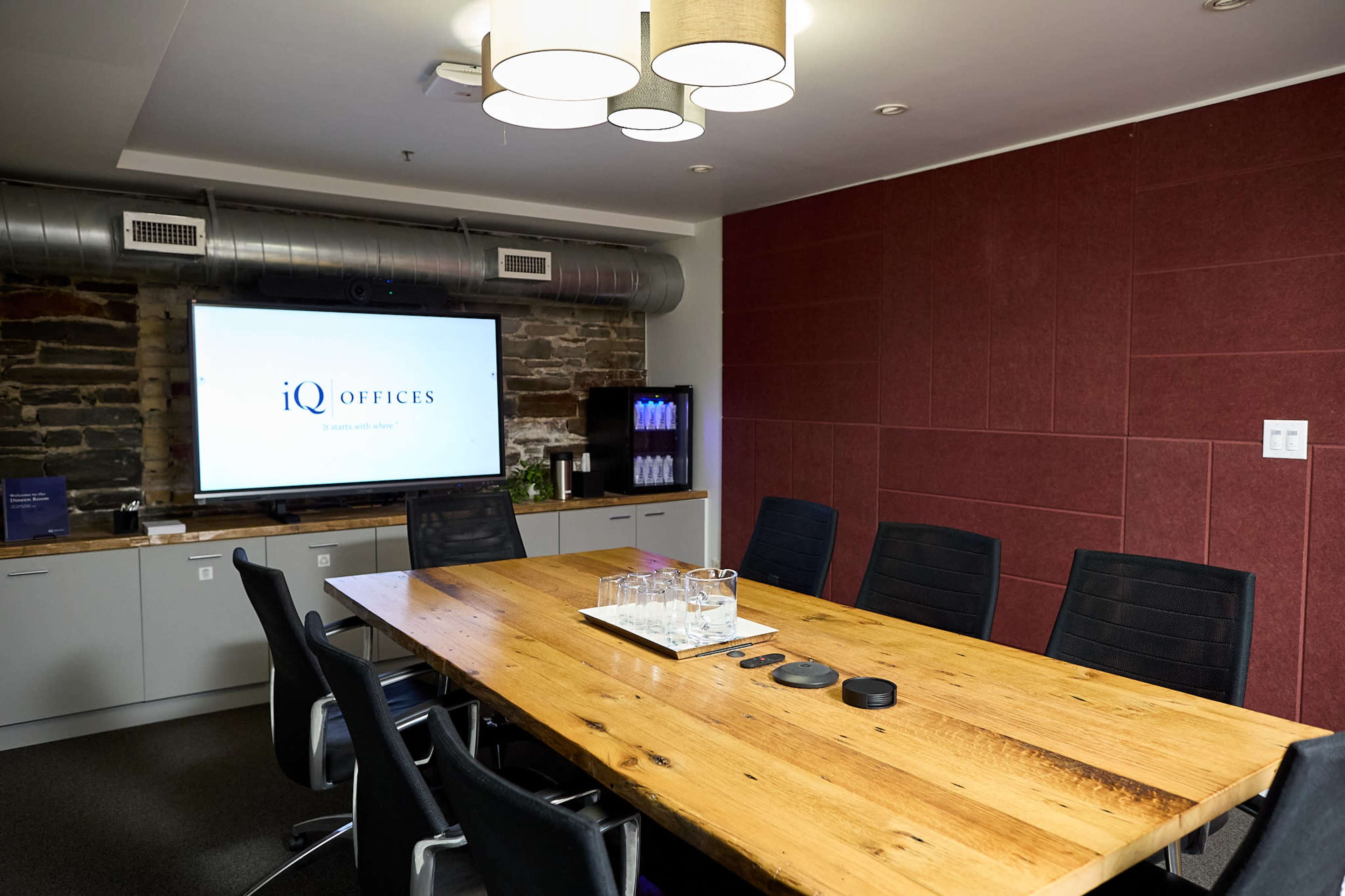 A modern conference room features a long wooden table surrounded by black chairs, a large screen mounted on the wall, and a side cabinet displaying water bottles.