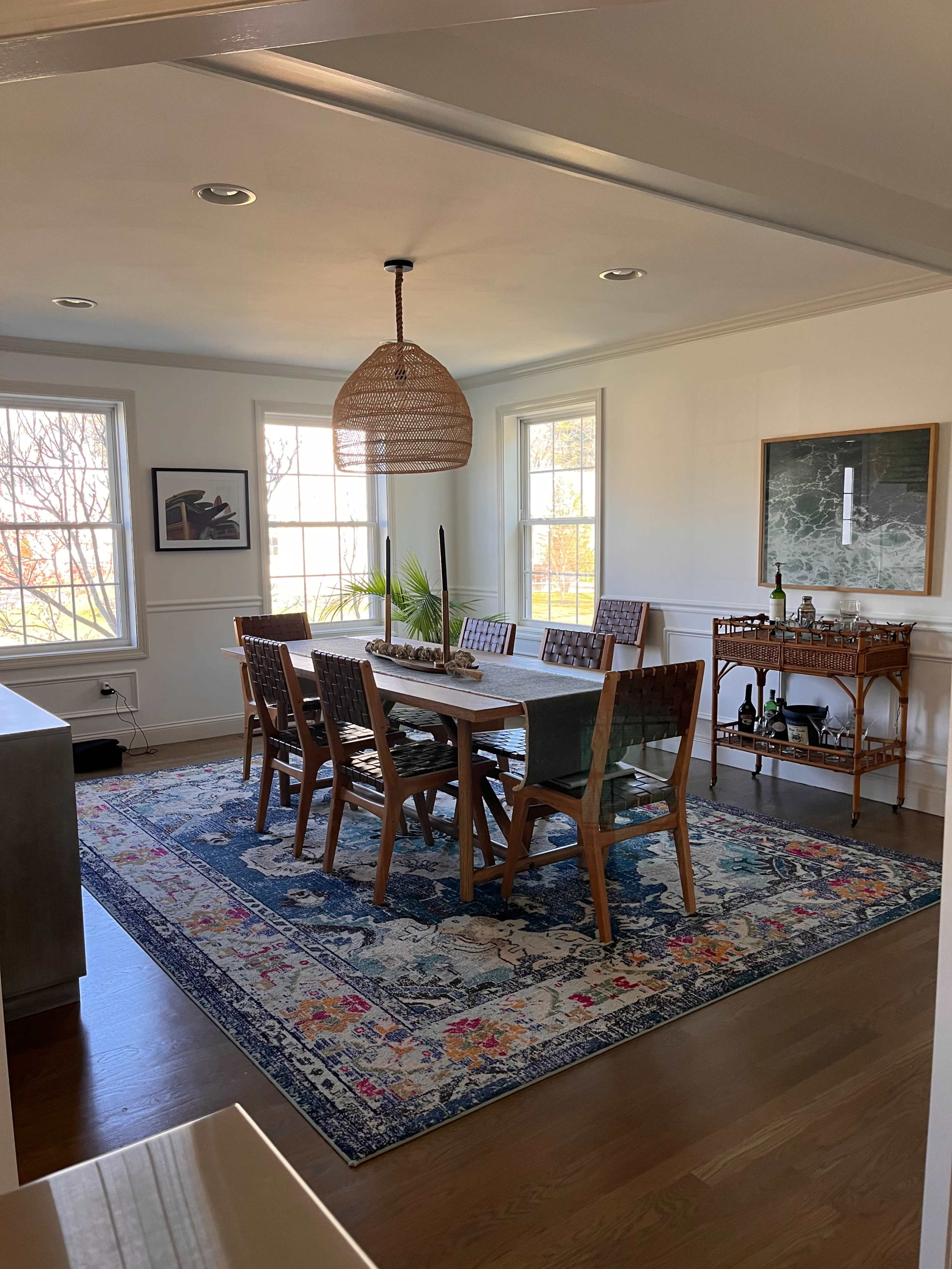 A dining room features a long wooden table with eight chairs, a large area rug, and windows allowing natural light.