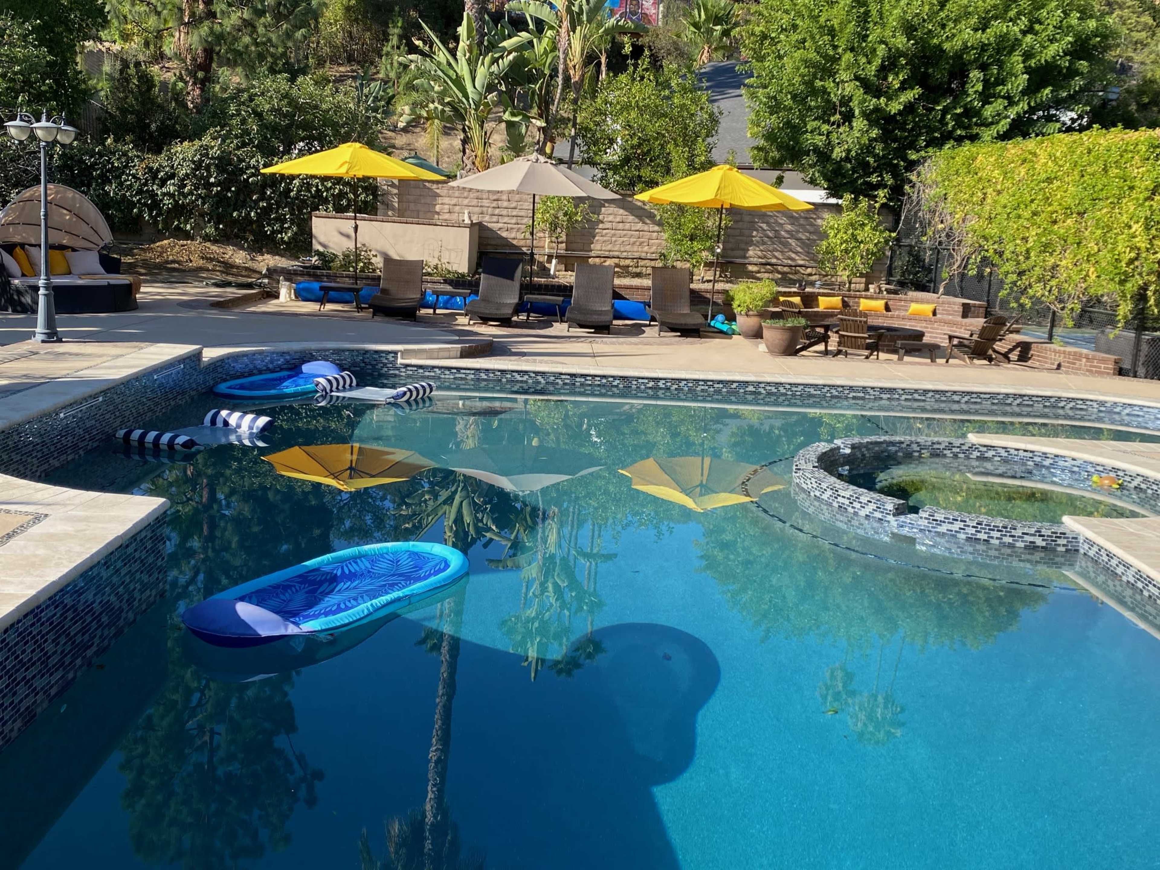 The image shows a swimming pool surrounded by yellow umbrellas, loungers, and inflatable floaties under clear blue skies.