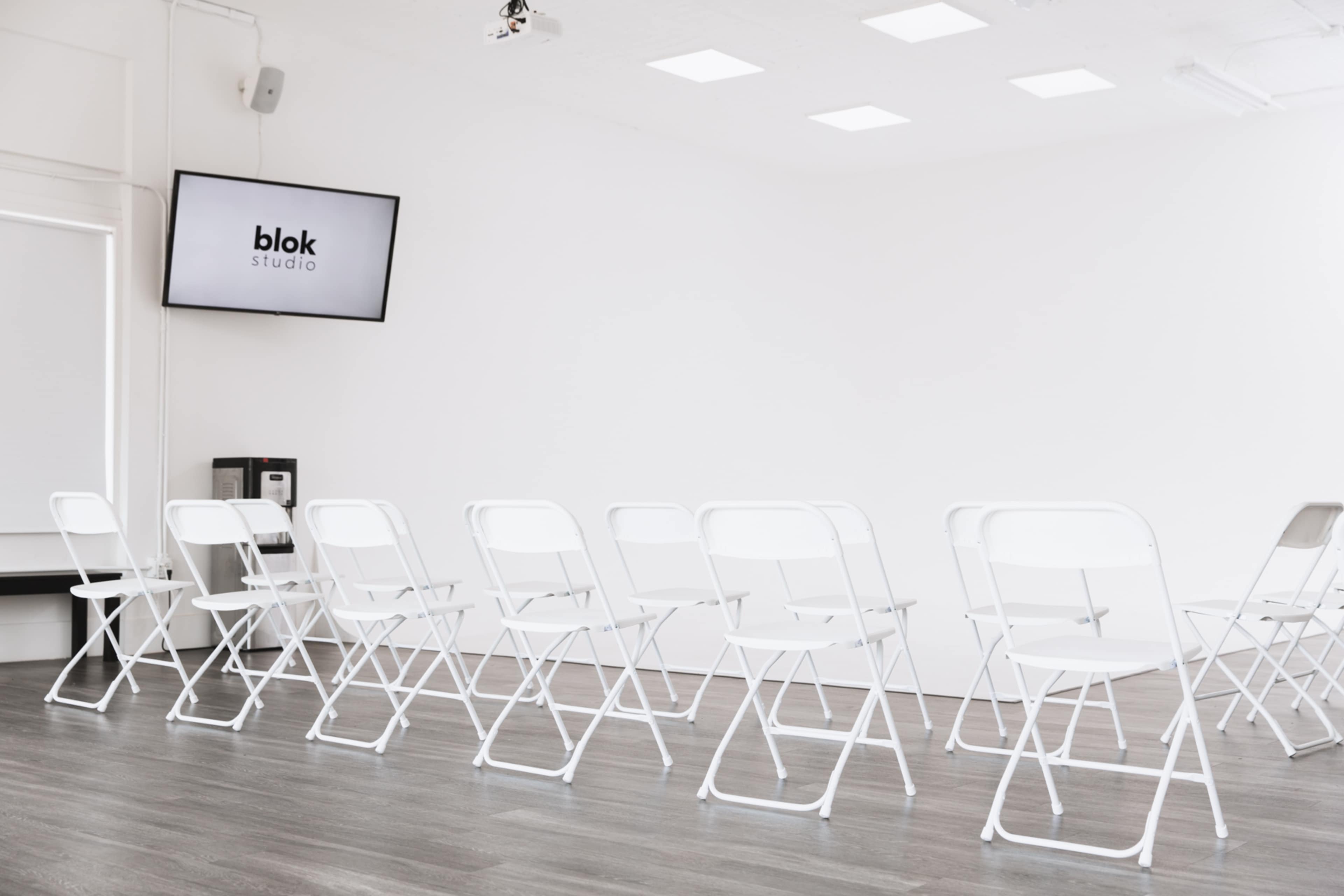 A minimalist studio space with white walls and a row of folding chairs facing a television screen displaying the logo "blok studio."