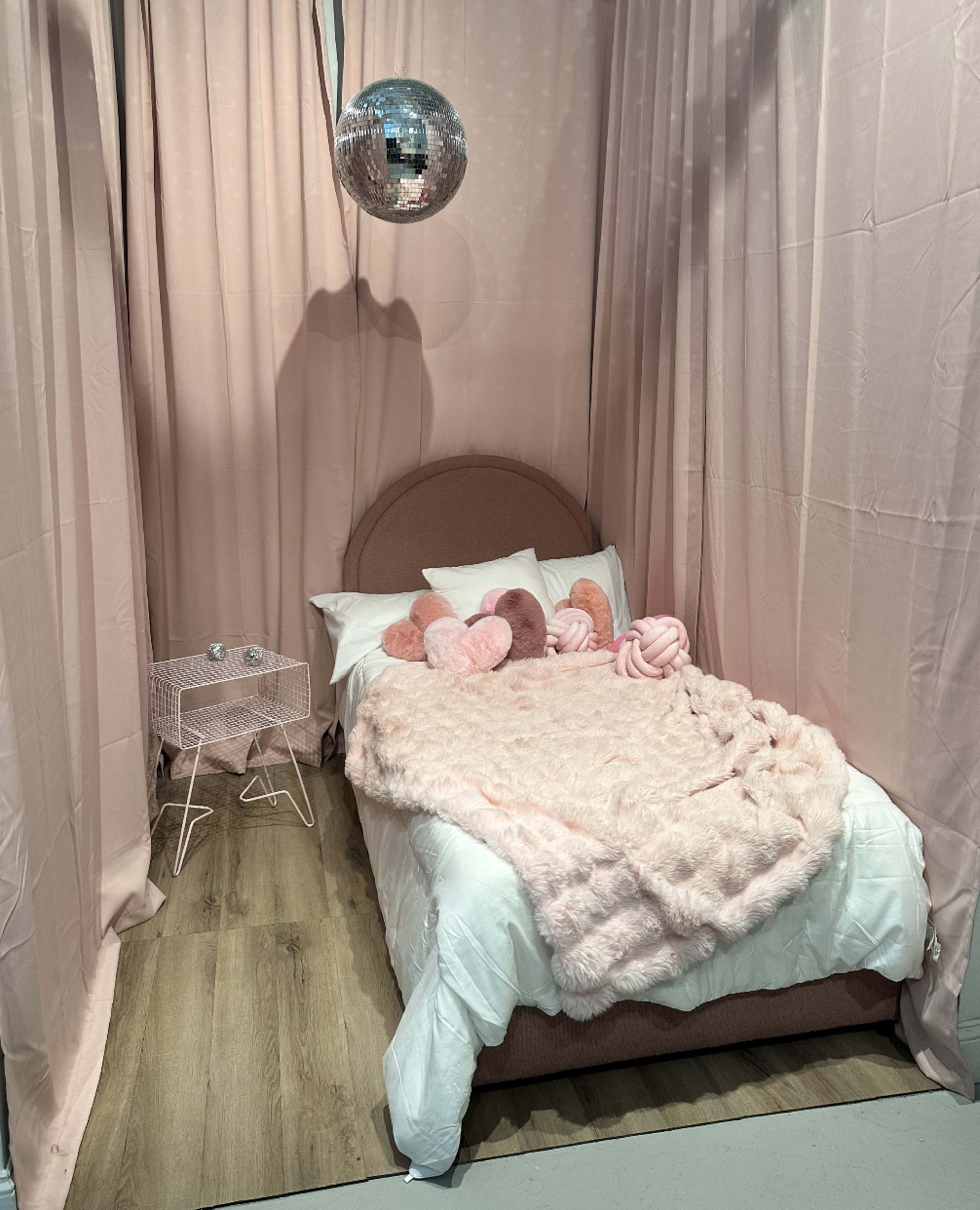 The image shows a neatly arranged bedroom with a pink bedspread, heart-shaped pillows, a disco ball hanging above, and pink curtains draping around the bed.