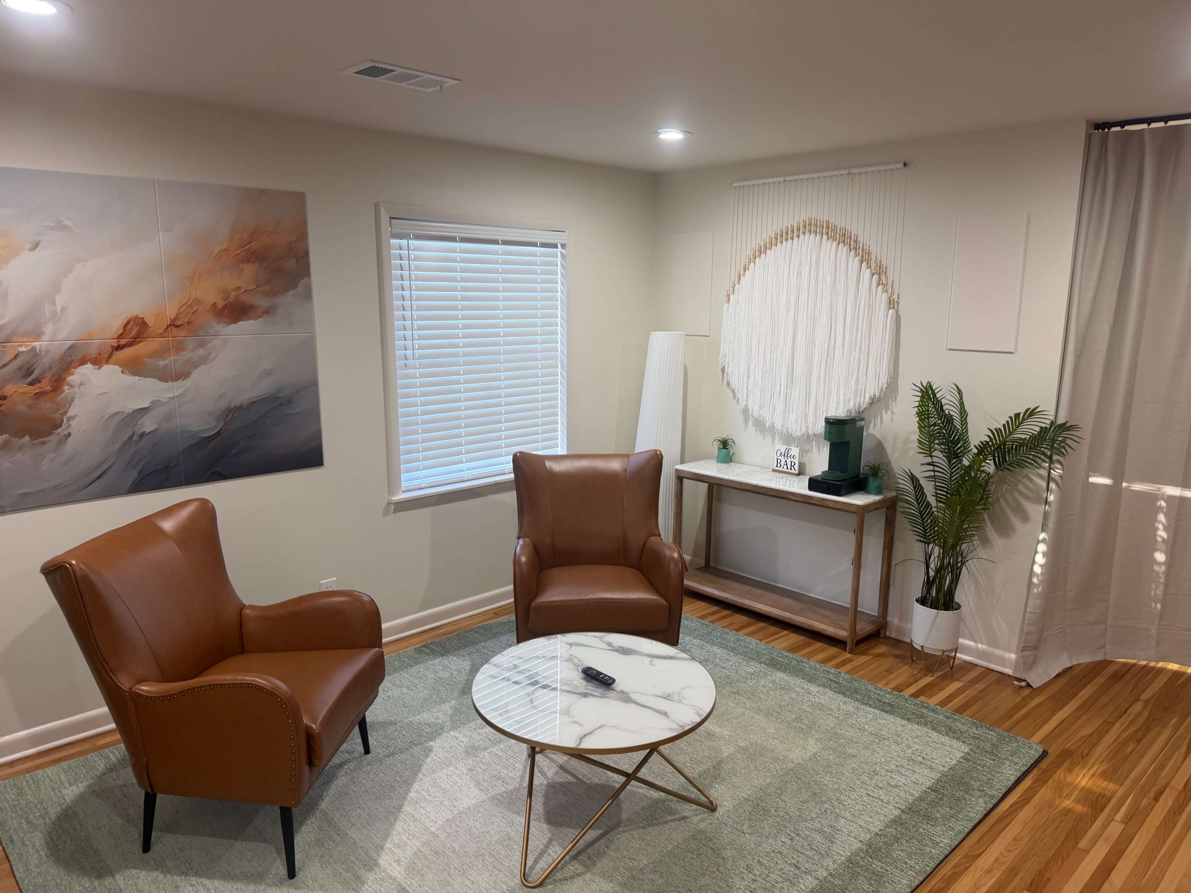 The image shows a cozy living room with two brown leather armchairs, a round marble coffee table, a potted plant, and a decorative wall hanging.