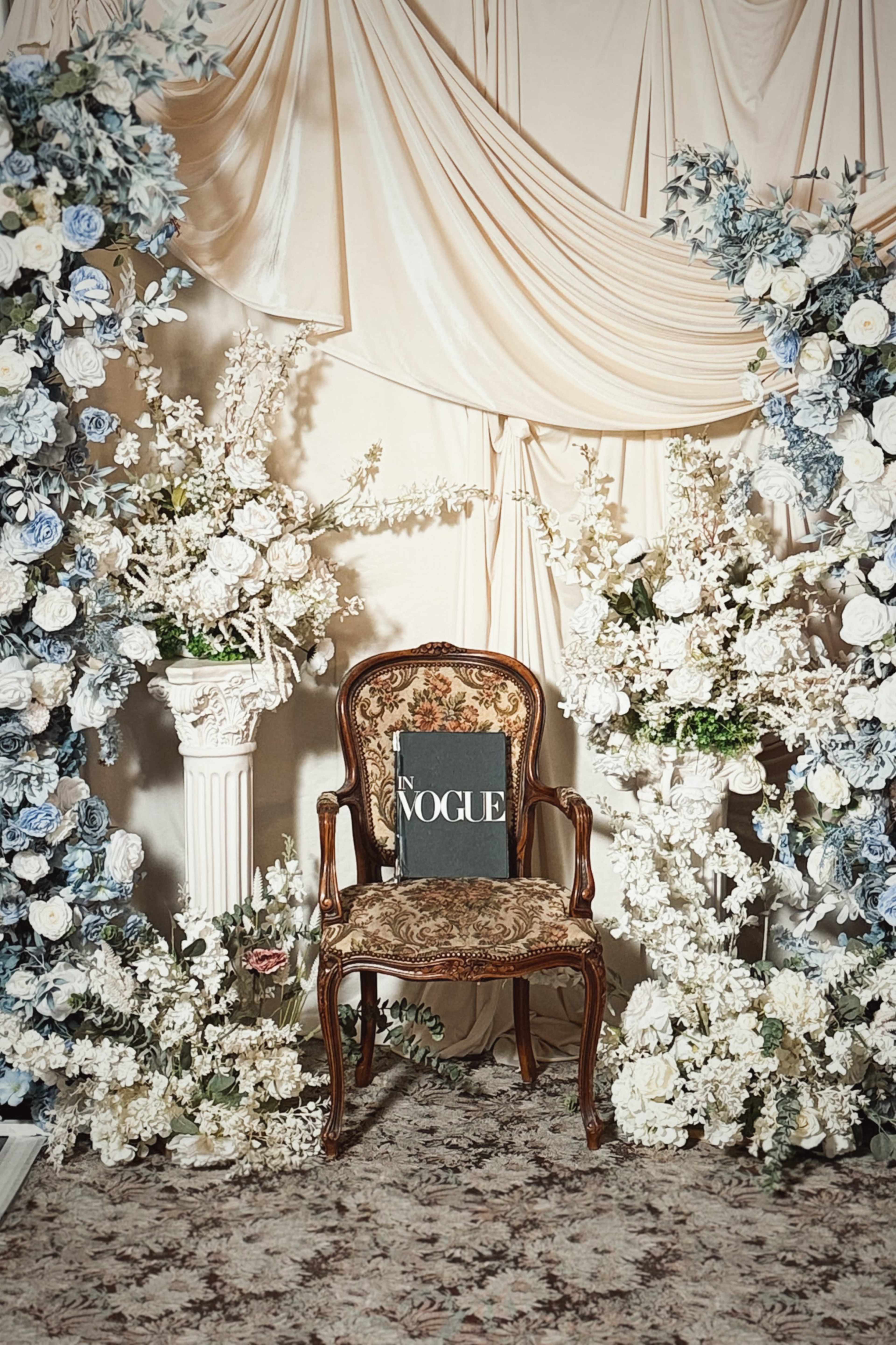 An ornate chair is positioned in front of a backdrop draped with fabric and adorned with floral arrangements and a copy of Vogue magazine resting on the seat.