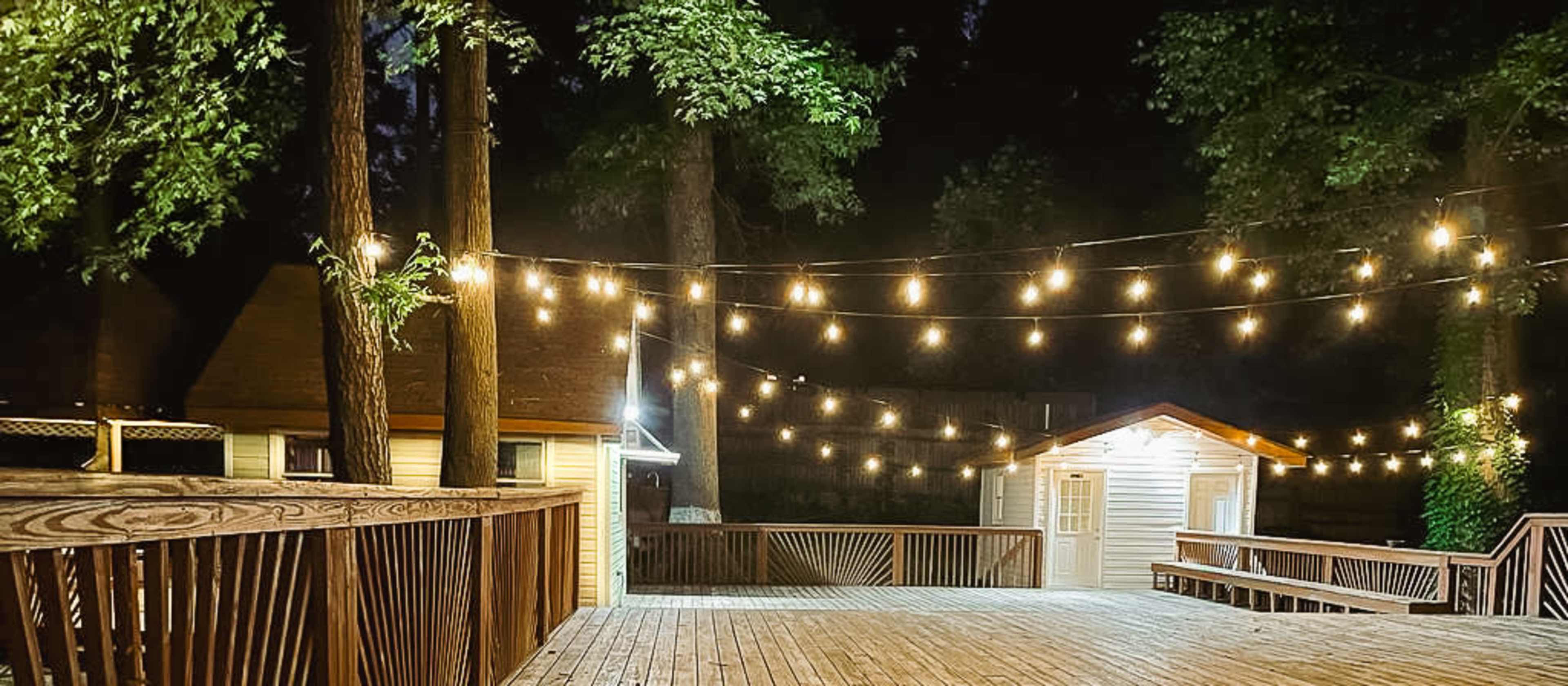 A wooden deck is illuminated at night by string lights, surrounded by trees and featuring a small building in the background.