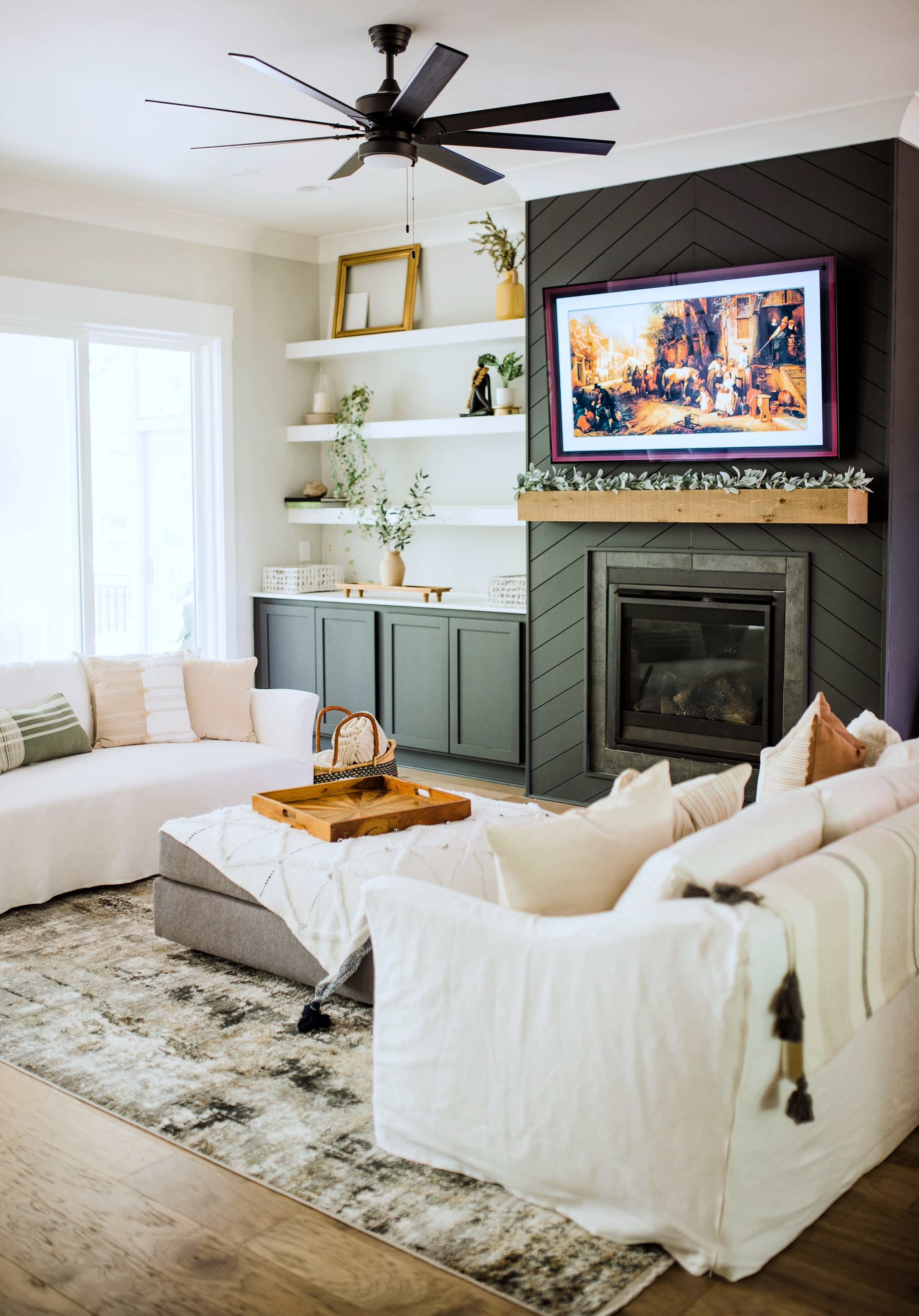 A modern living room features a ceiling fan, a fireplace with a television above it, and a sectional sofa arranged around a coffee table on a patterned rug.