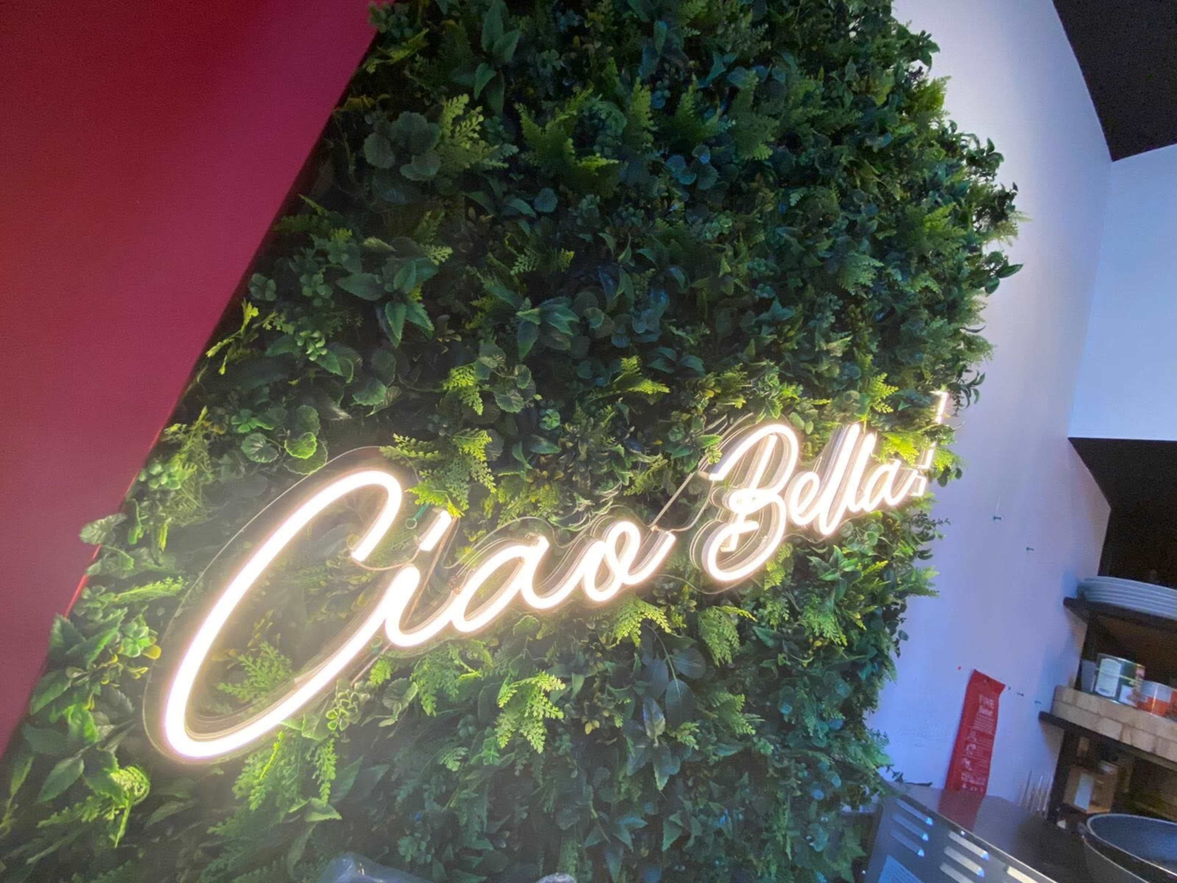 A neon sign that says "Ciao Bellas!" is illuminated against a backdrop of green foliage.