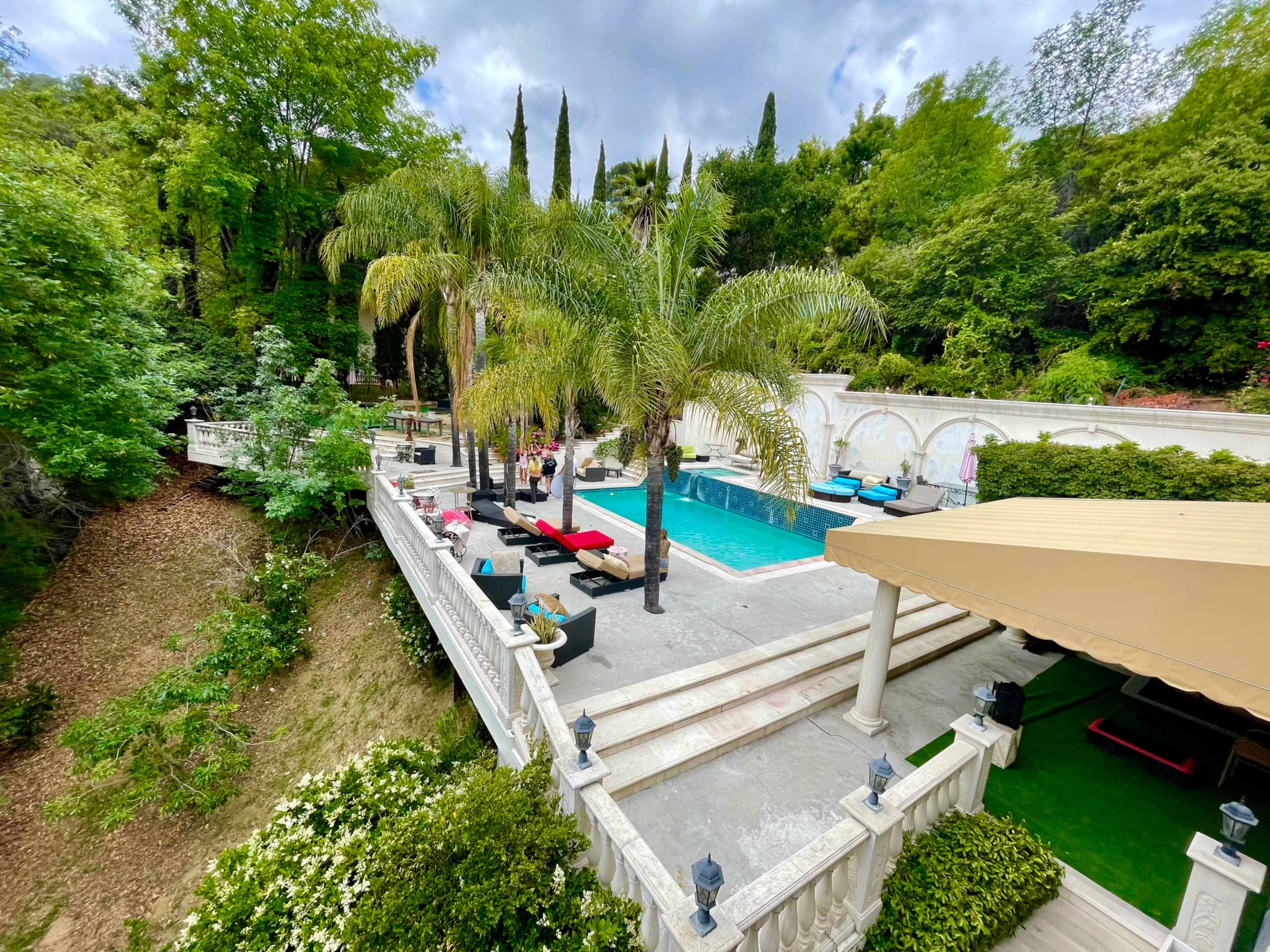 A swimming pool area surrounded by palm trees and lush greenery, with lounge chairs and a terrace.