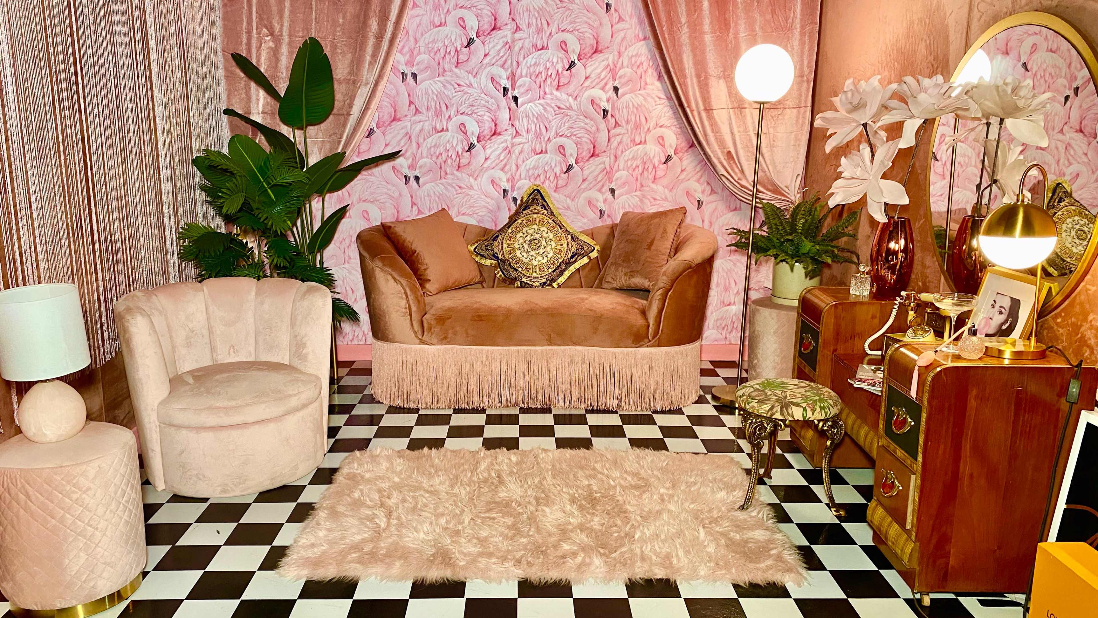 A softly lit lounge area features a pink velvet sofa, a round chair, a black and white checkered floor, and a decorative wall with a flamingo pattern.
