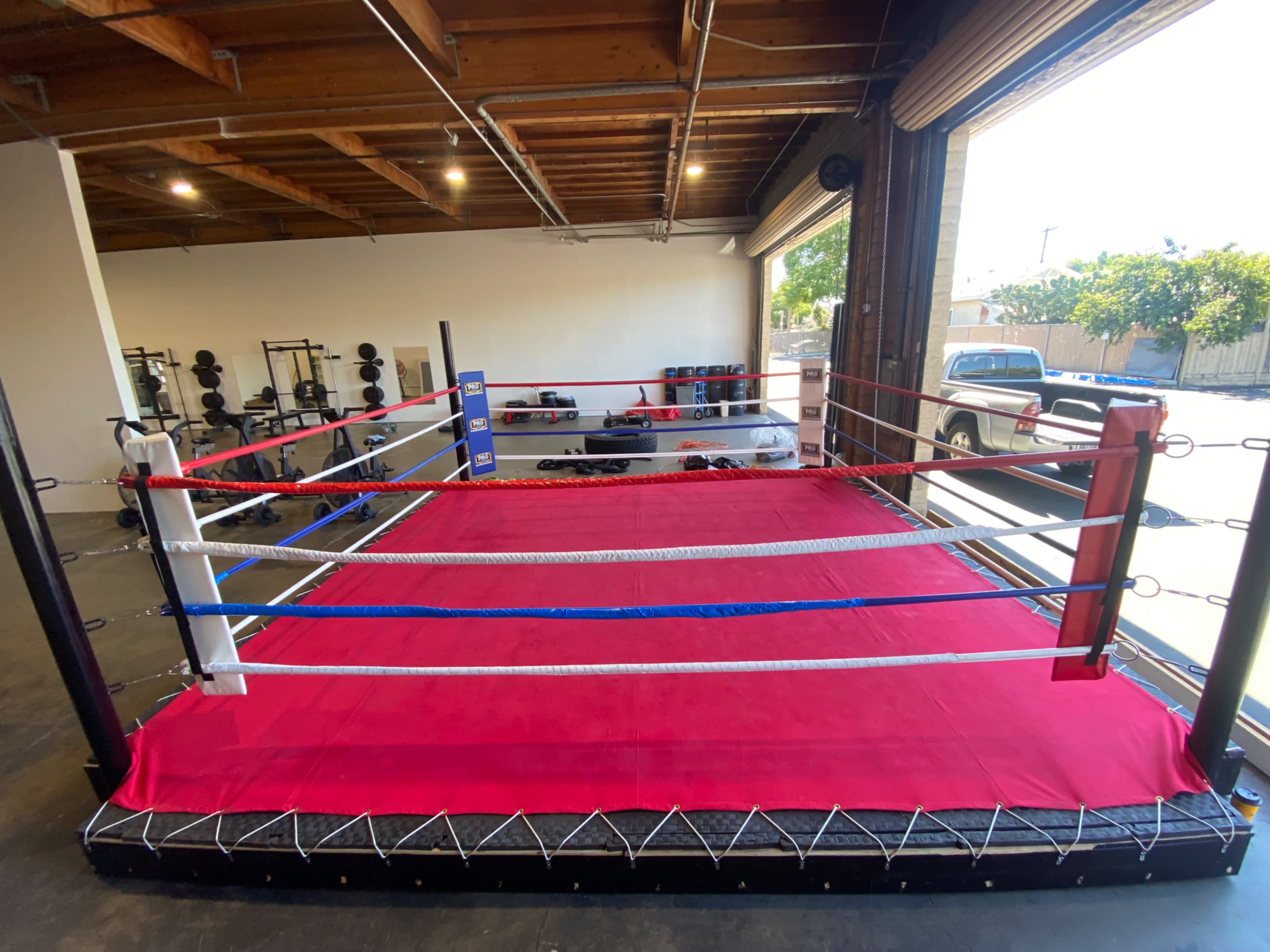A boxing ring with red and blue corner pads set up in a gym with weightlifting equipment in the background.