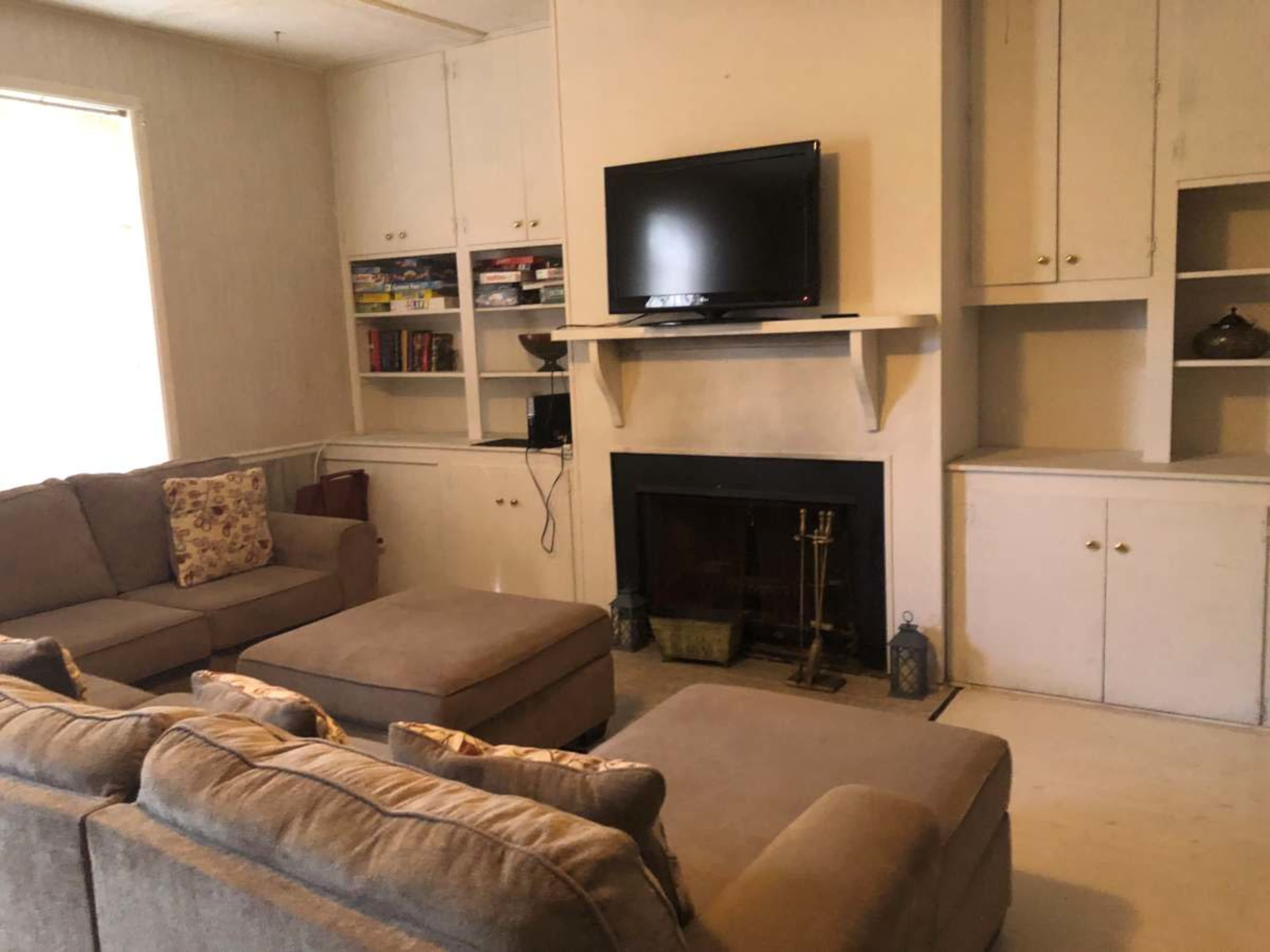 A living room features a large sectional couch, a flat-screen television mounted above a fireplace, and built-in shelves filled with books and decor.