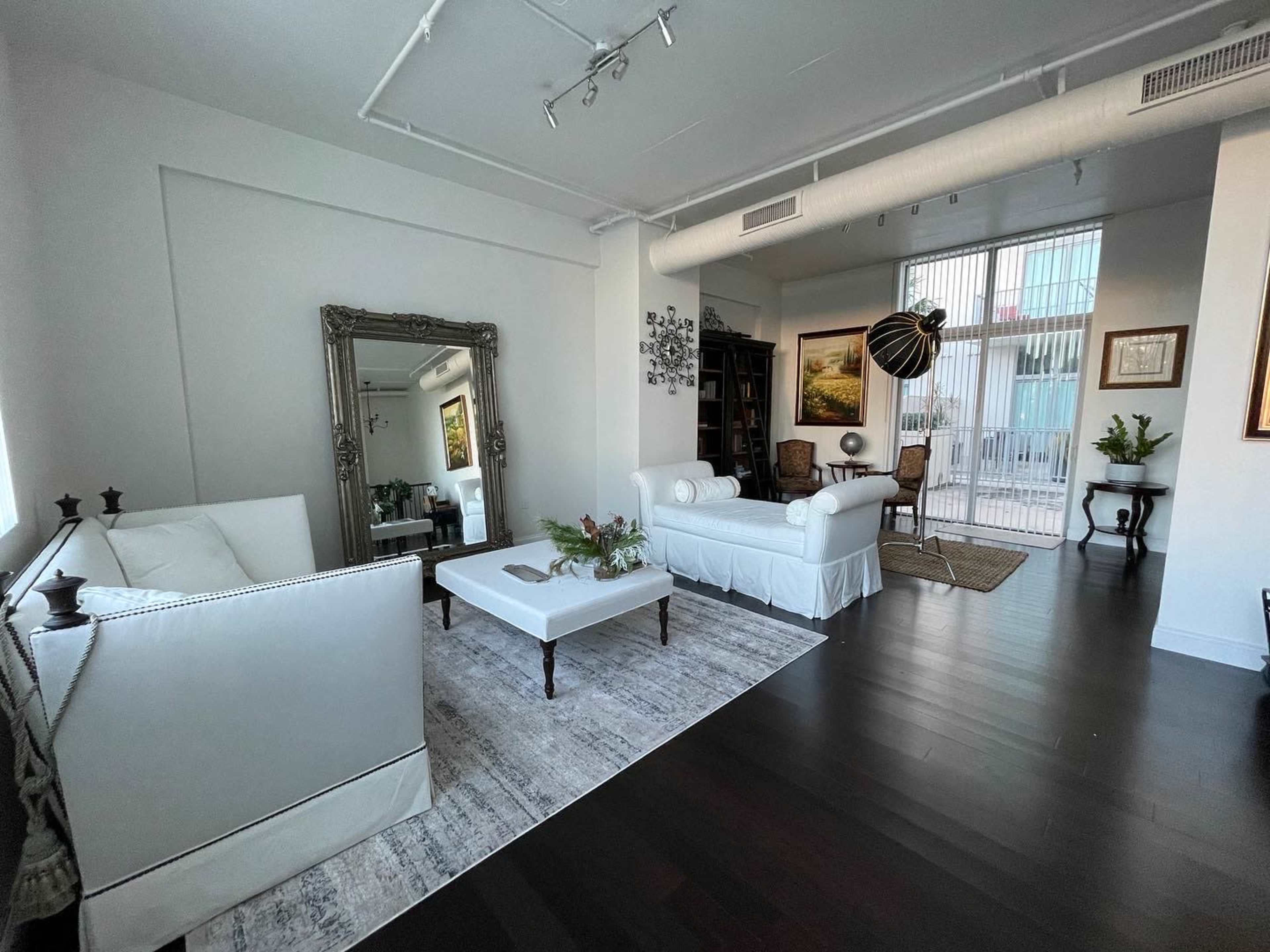 The image shows a contemporary living room with a white sofa, a coffee table with flowers, and large windows revealing an outdoor balcony.