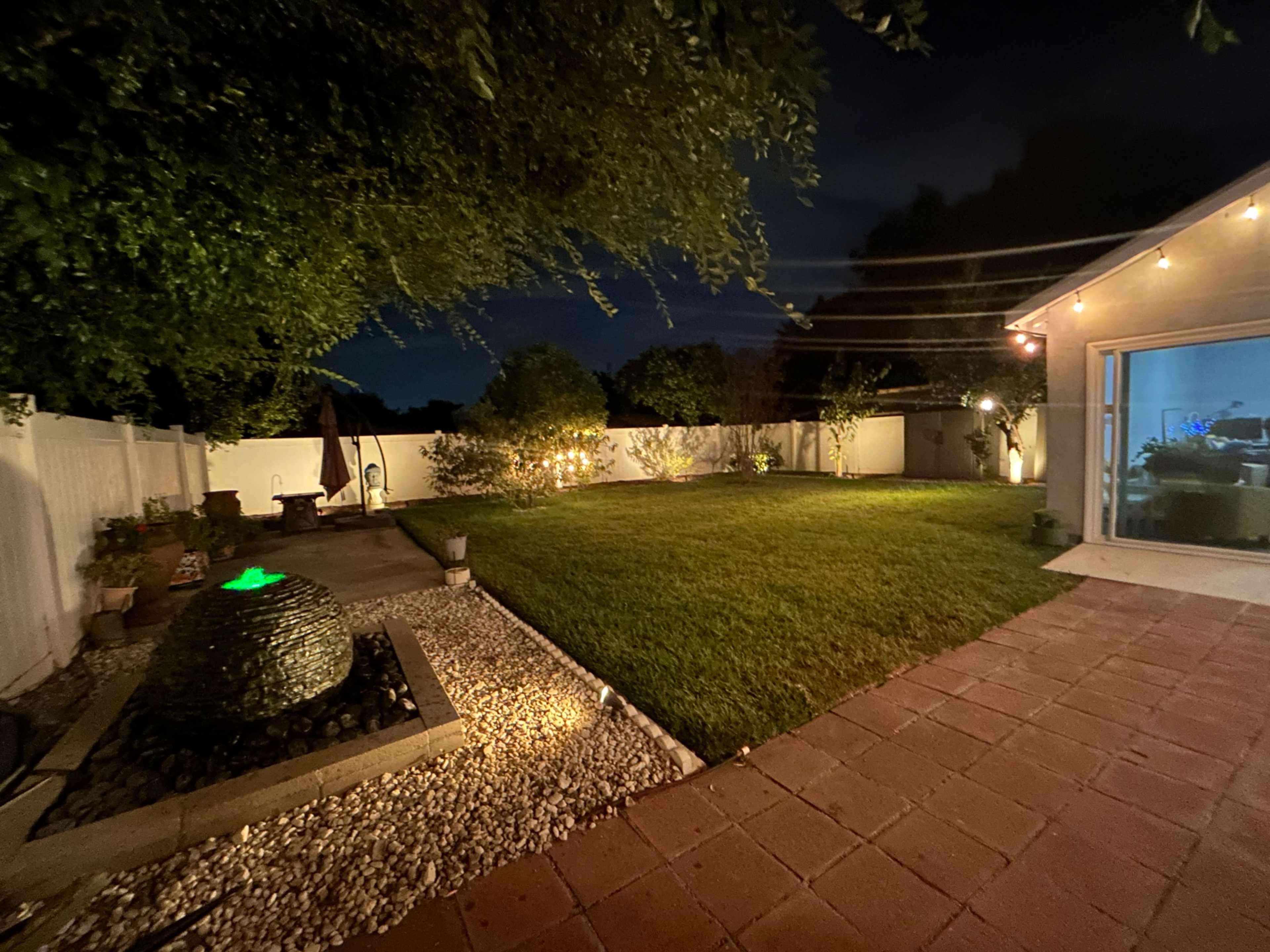 The image shows a backyard at night with a green-lit water fountain and a lawn bordered by a white fence and patio area.