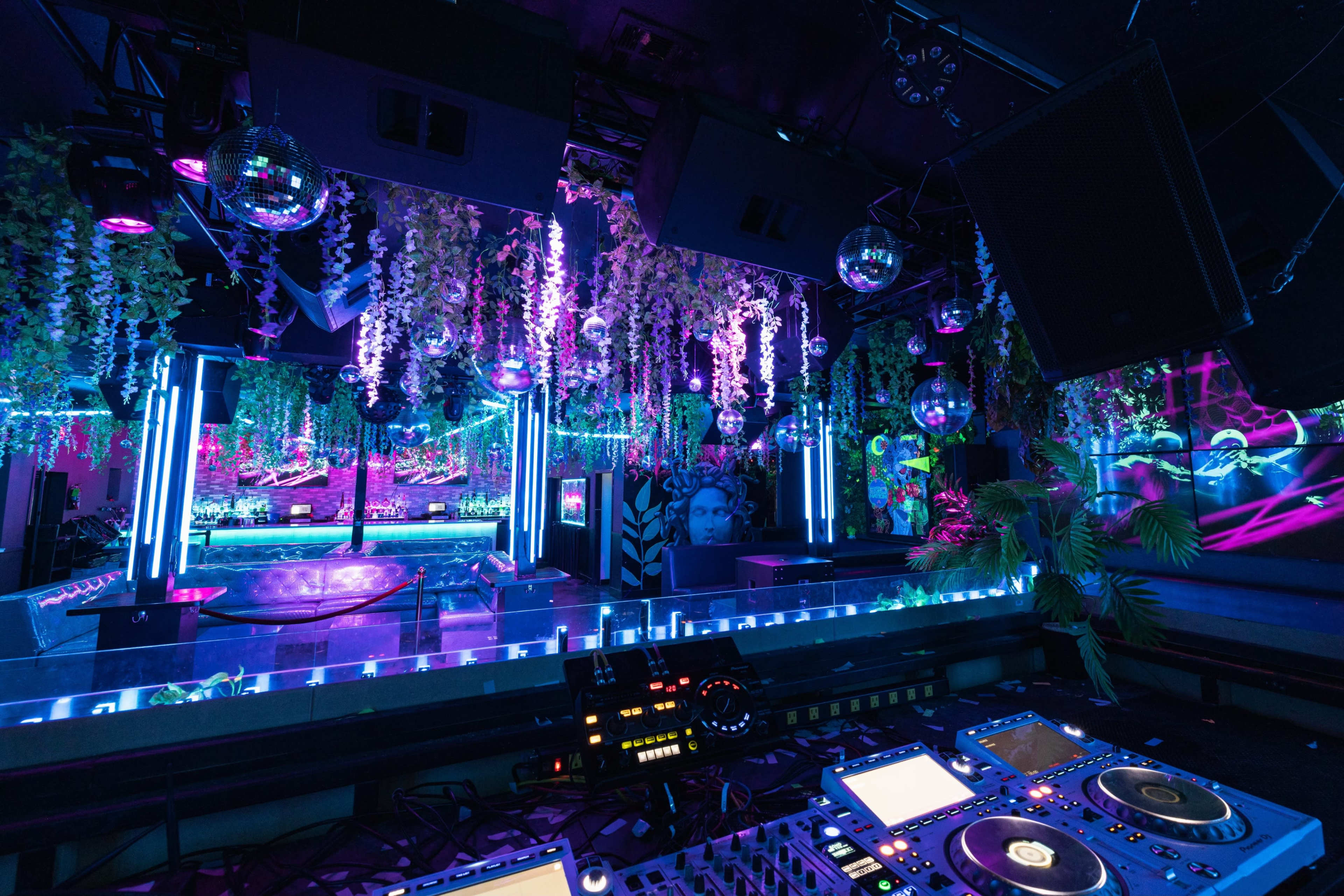 A vibrant nightclub interior features colorful lighting, decorative plants, and disco balls above a DJ booth.