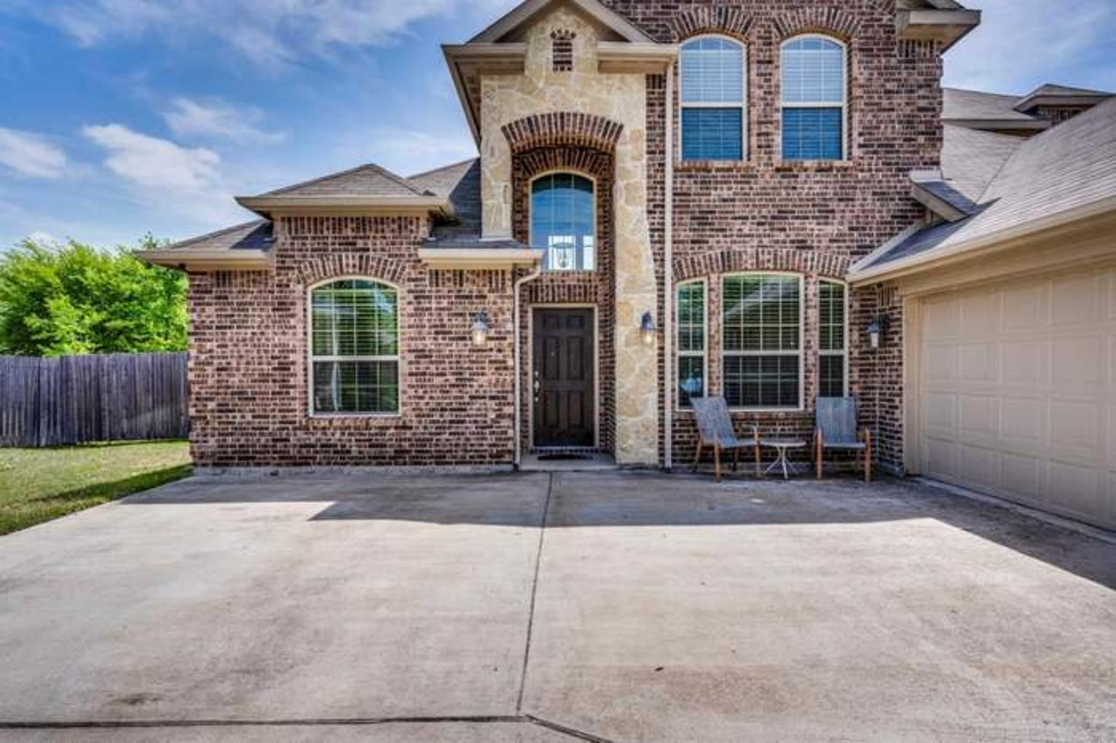 The image shows the front exterior of a two-story brick house with large windows and a driveway leading to a garage.