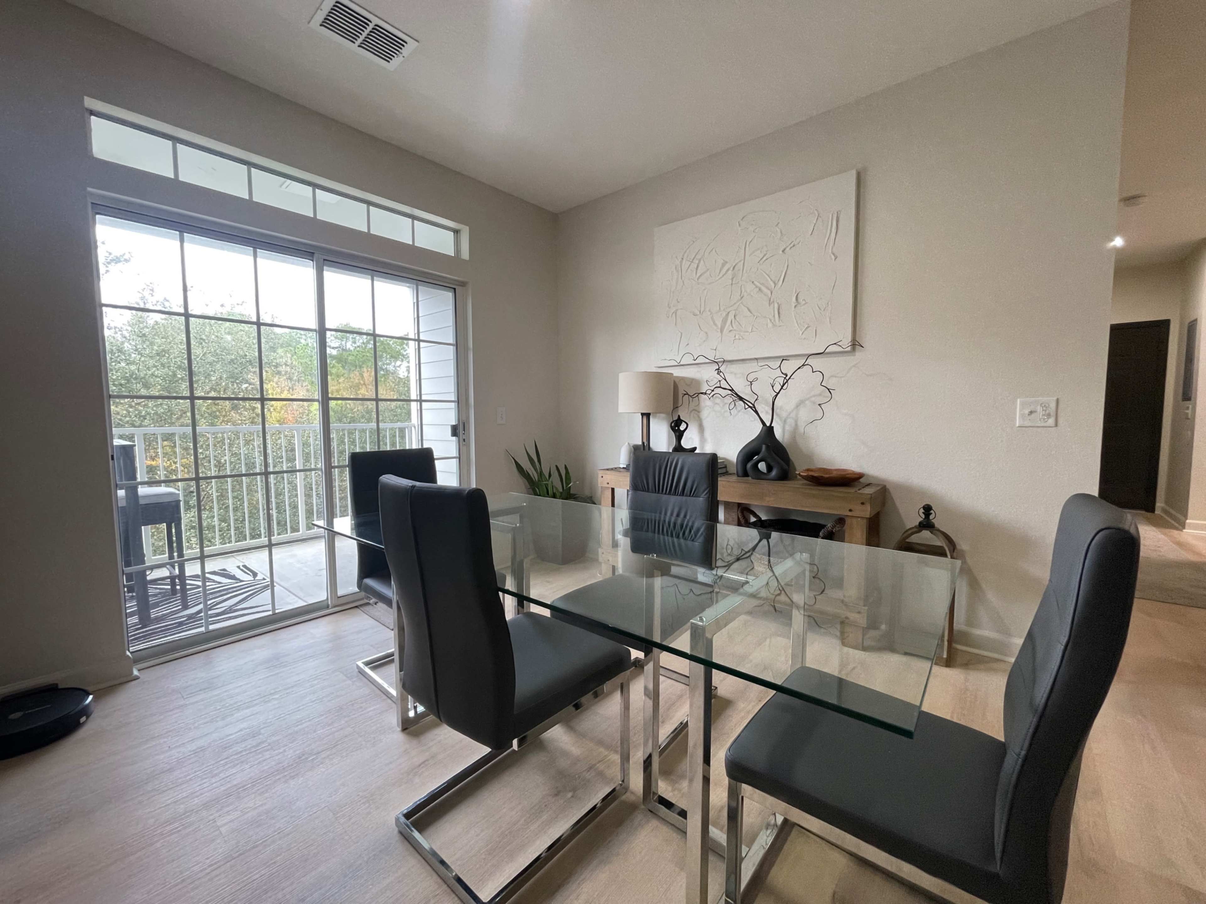 A glass dining table with four chairs is positioned near a sliding door that opens to a balcony, complemented by a minimalistic decor in a well-lit room.