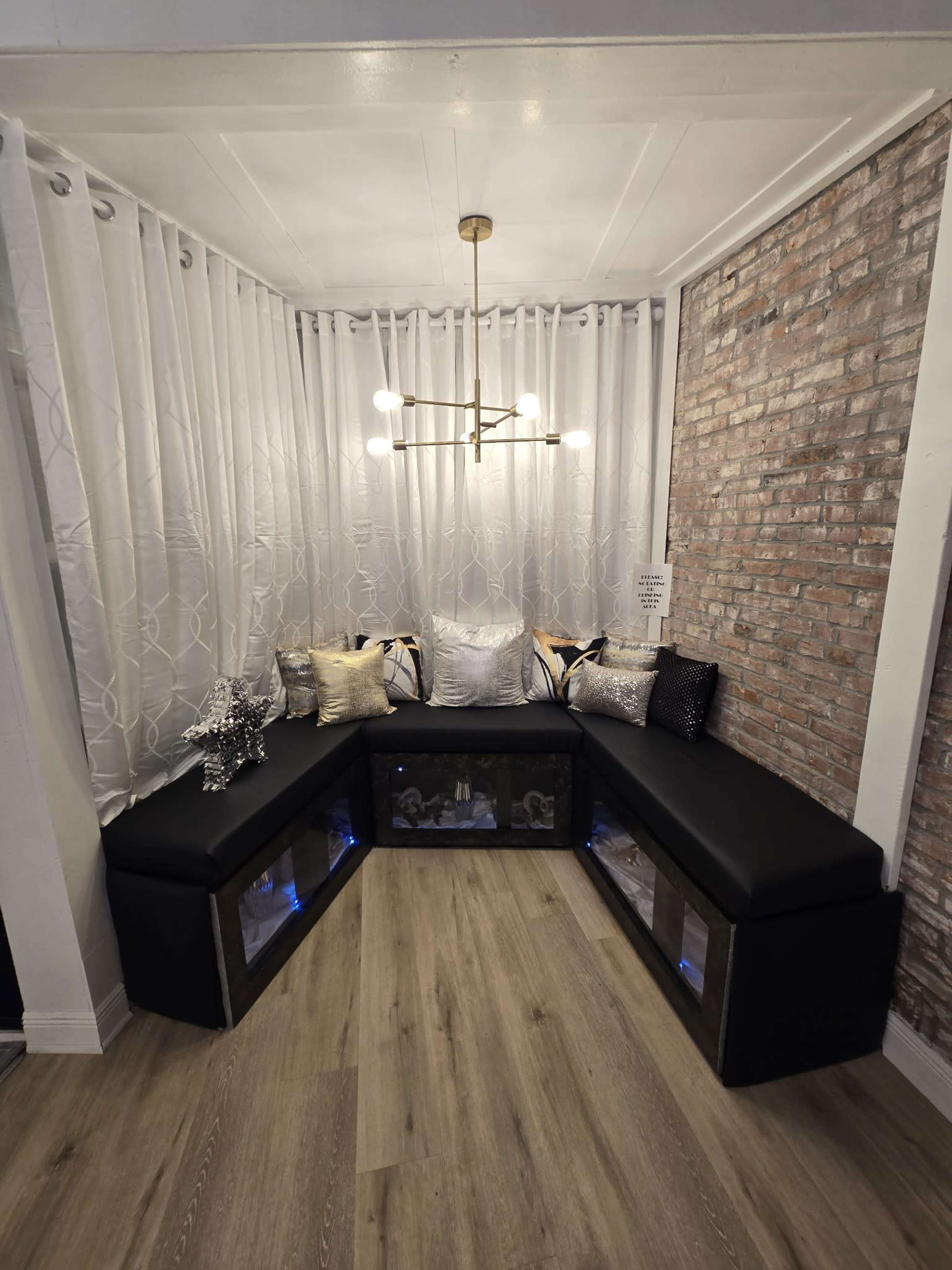 The image shows a modern corner seating area with a black upholstered bench, decorative pillows, sheer white curtains, a brick wall, and a contemporary light fixture.
