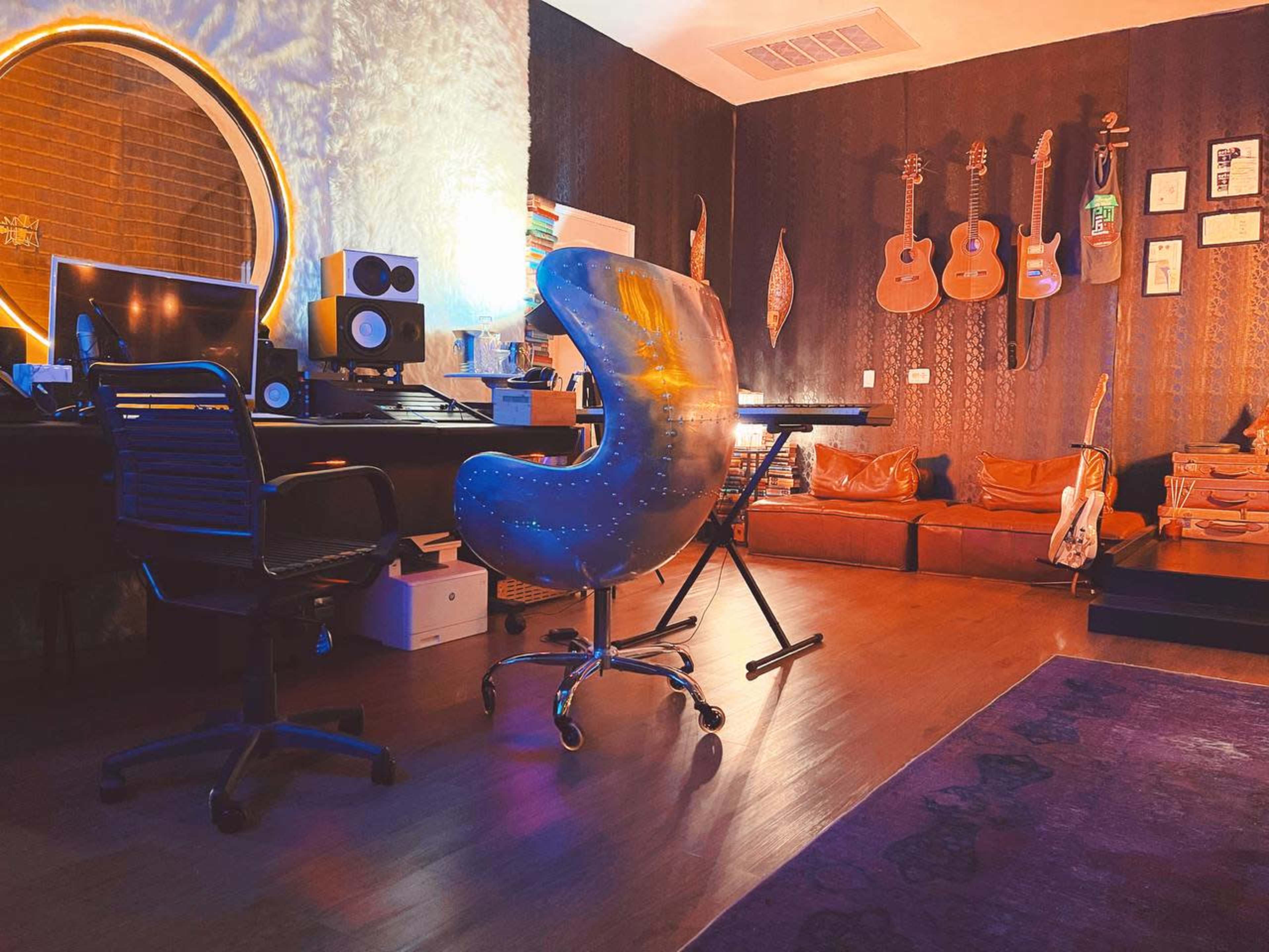 A music studio features a modern ergonomic chair facing a console with audio equipment, surrounded by guitars hanging on the wall and a leather couch.