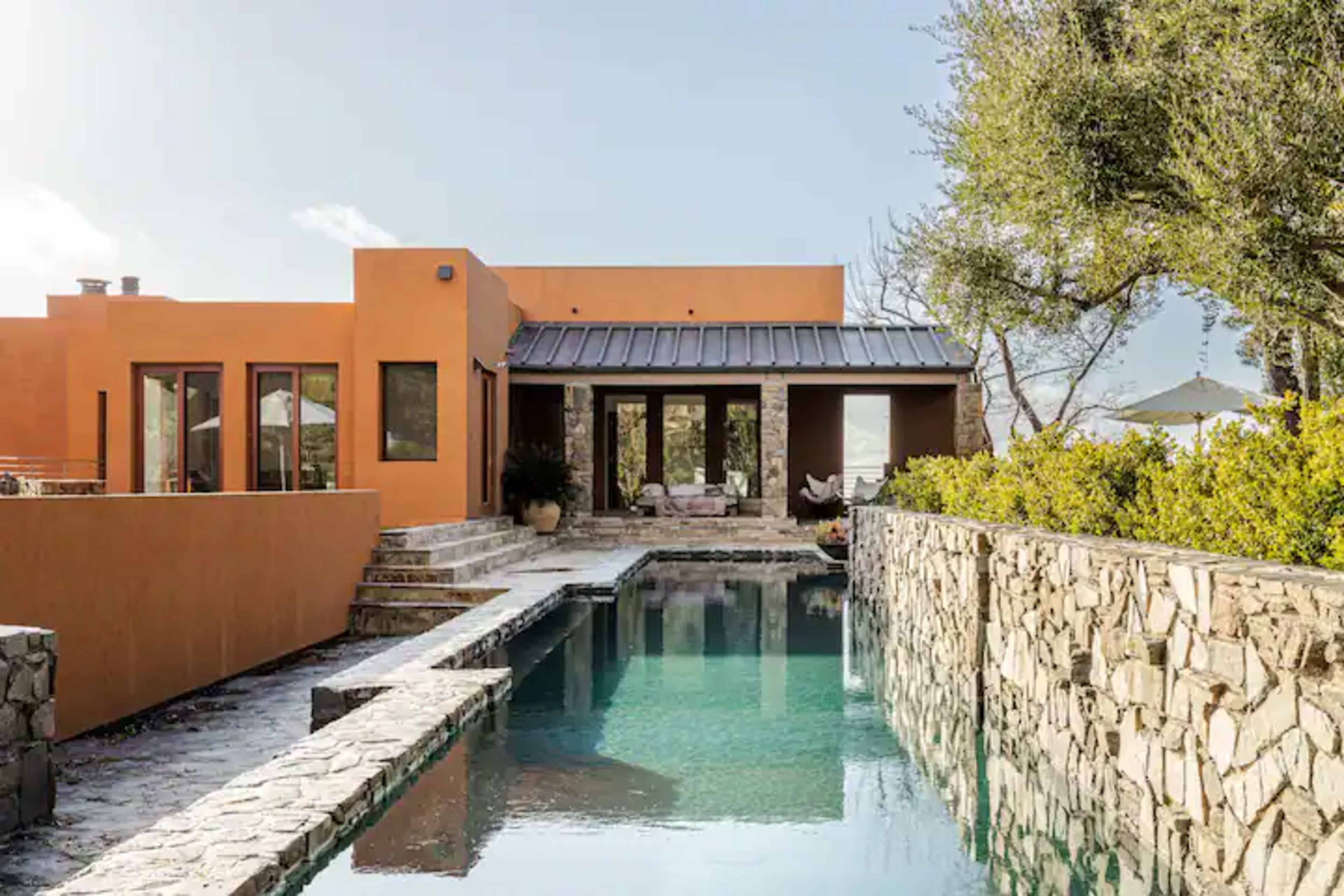 A modern, orange stucco house with large windows overlooks a reflective pool and stone landscaping.