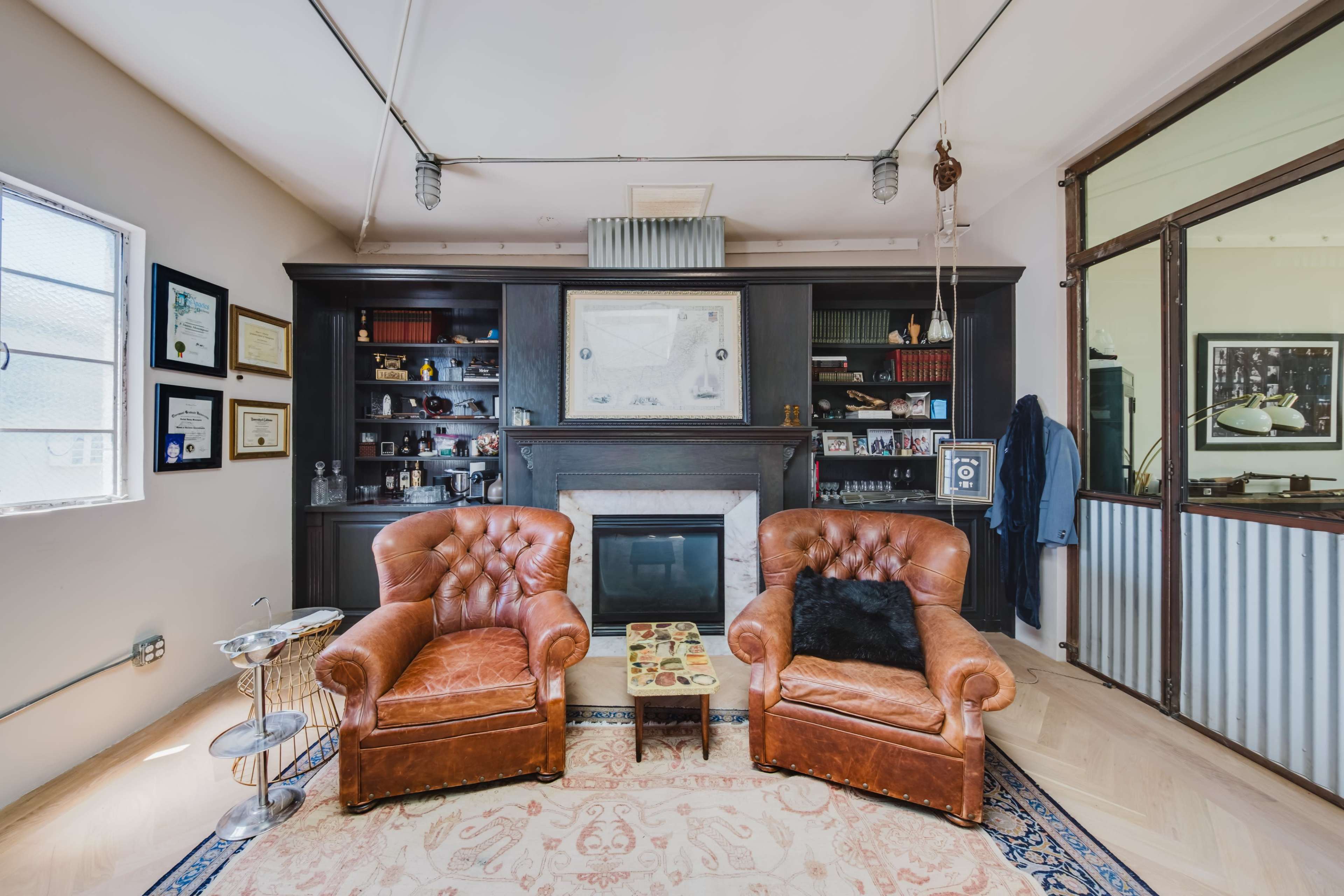 The image shows a living room with two brown leather armchairs, a fireplace, and a bookshelf filled with various items and framed certificates, set against a light wooden floor.