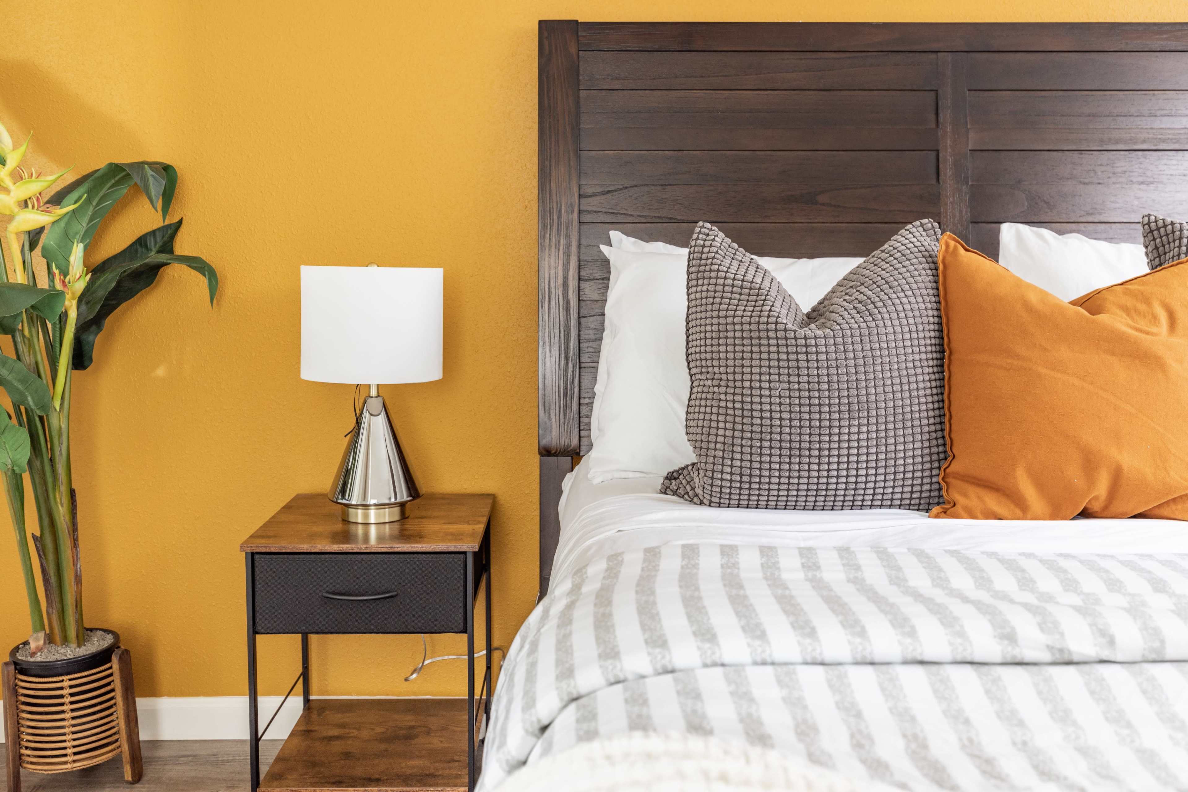 A bed with decorative pillows is positioned next to a wooden nightstand holding a lamp, against a bright orange wall.