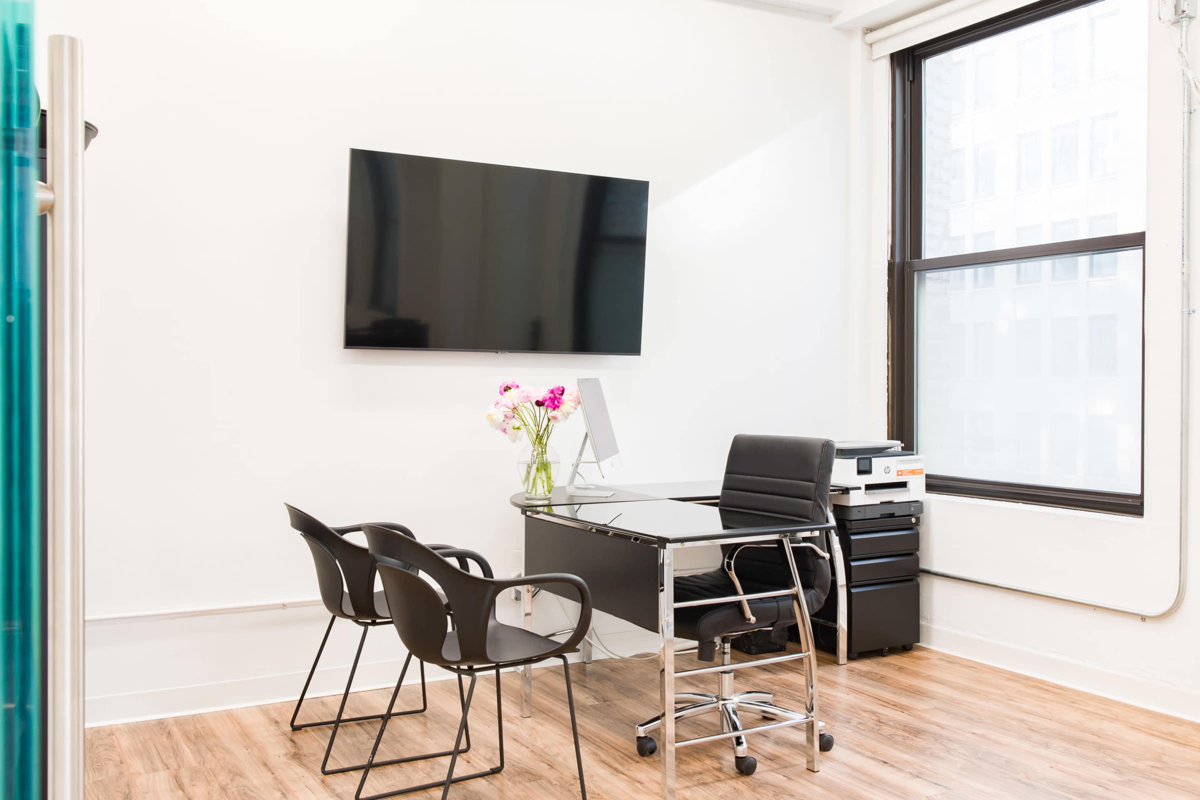 A modern office space features a desk, three chairs, a computer, a printer, a television mounted on the wall, and a vase of flowers.