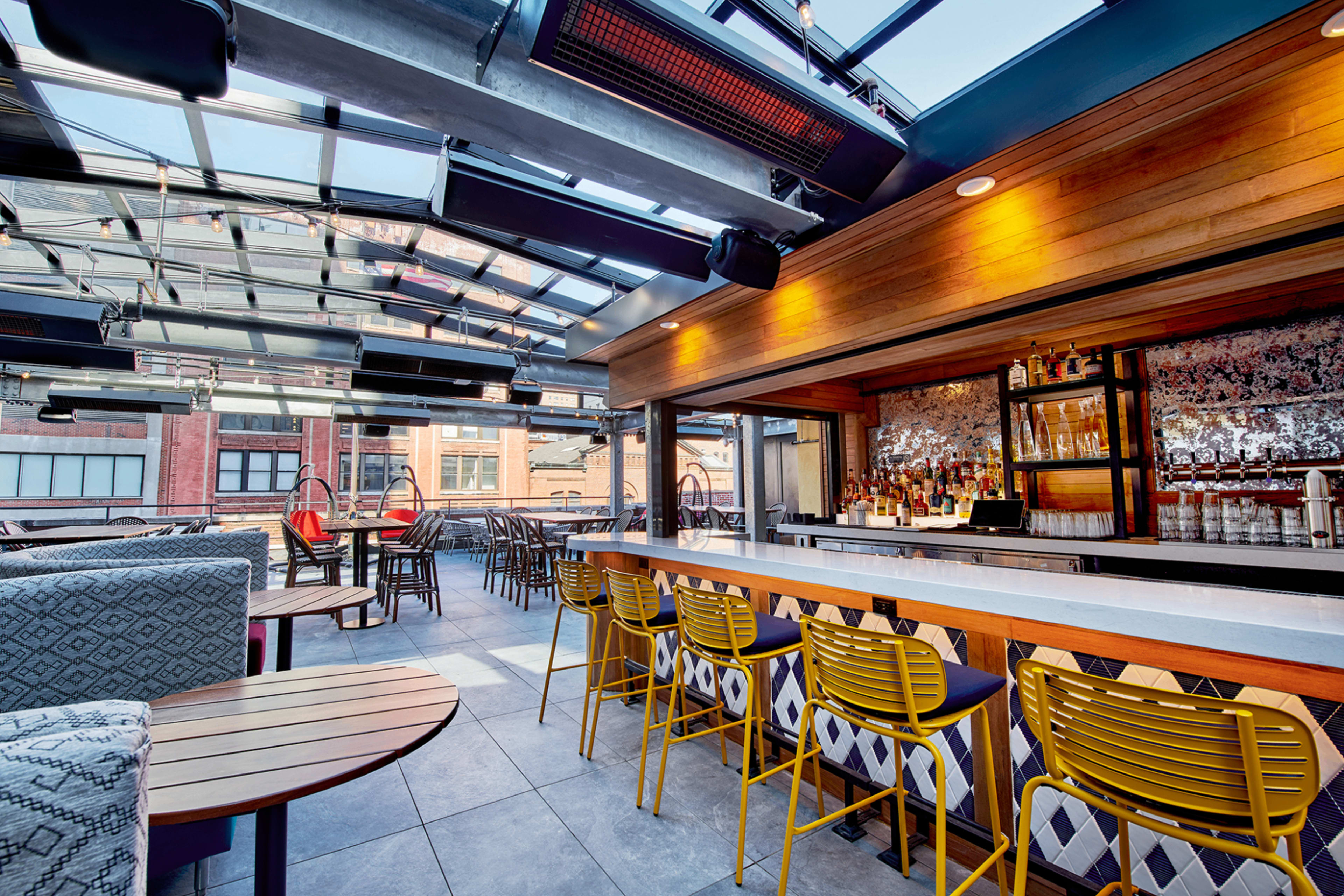 The image shows a modern rooftop bar with a glass ceiling, featuring wooden and metal seating arrangements and a well-stocked bar in the background.