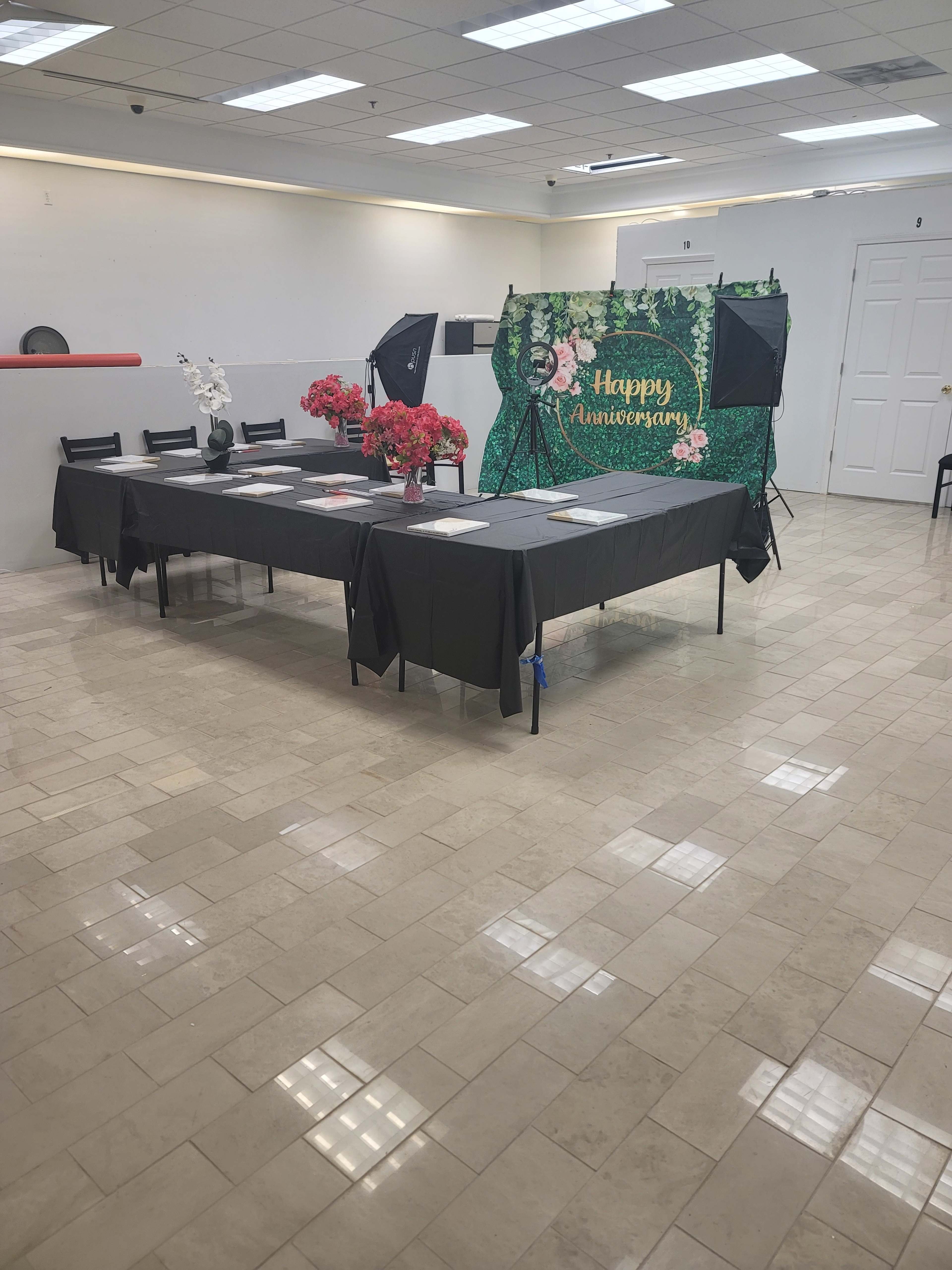 A large, empty room features a table set with black tablecloths, decorations of flowers, and a backdrop that reads "Happy Anniversary."