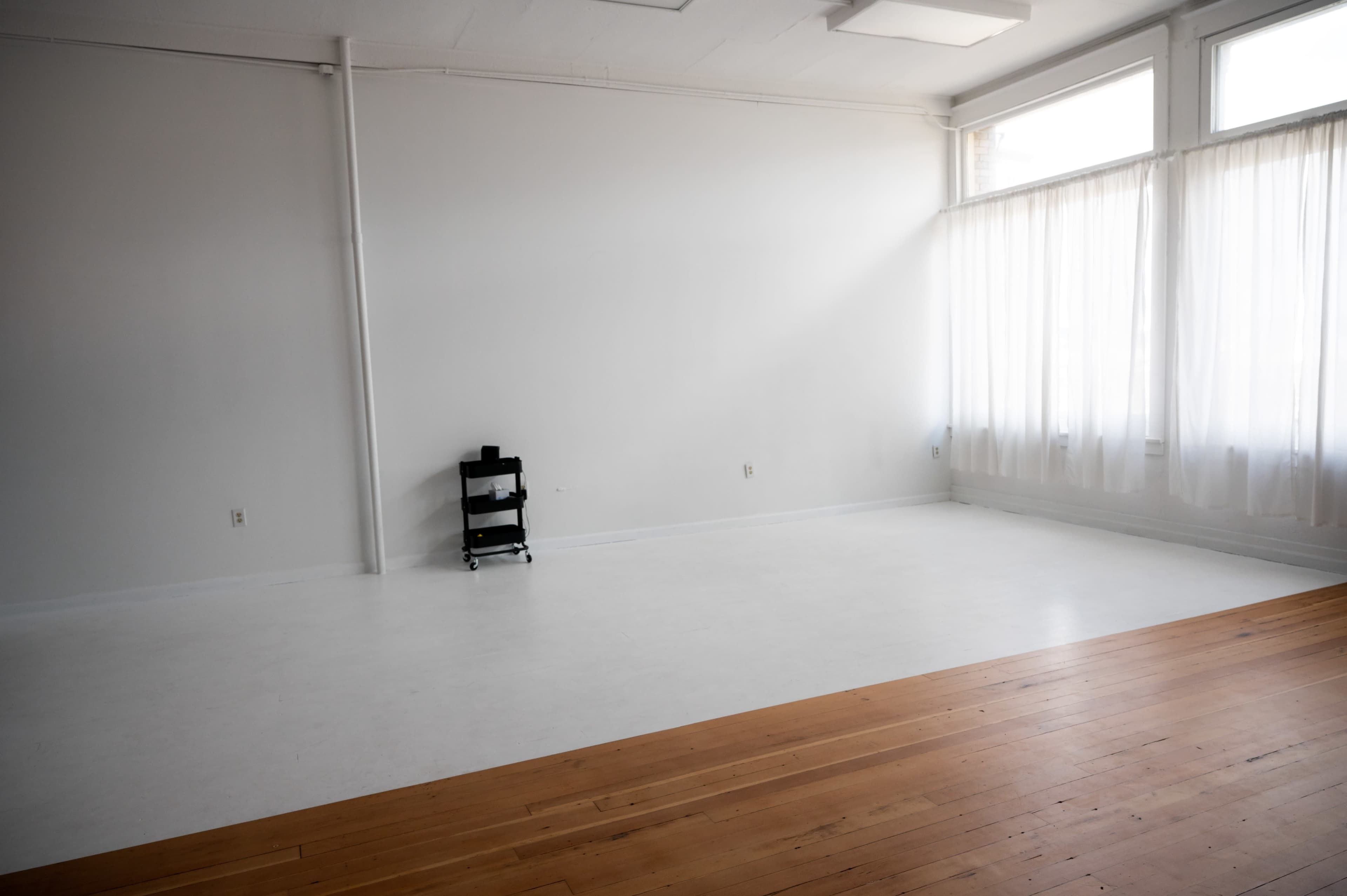 A spacious, empty room with white walls, a wooden floor, and large windows covered by sheer curtains.