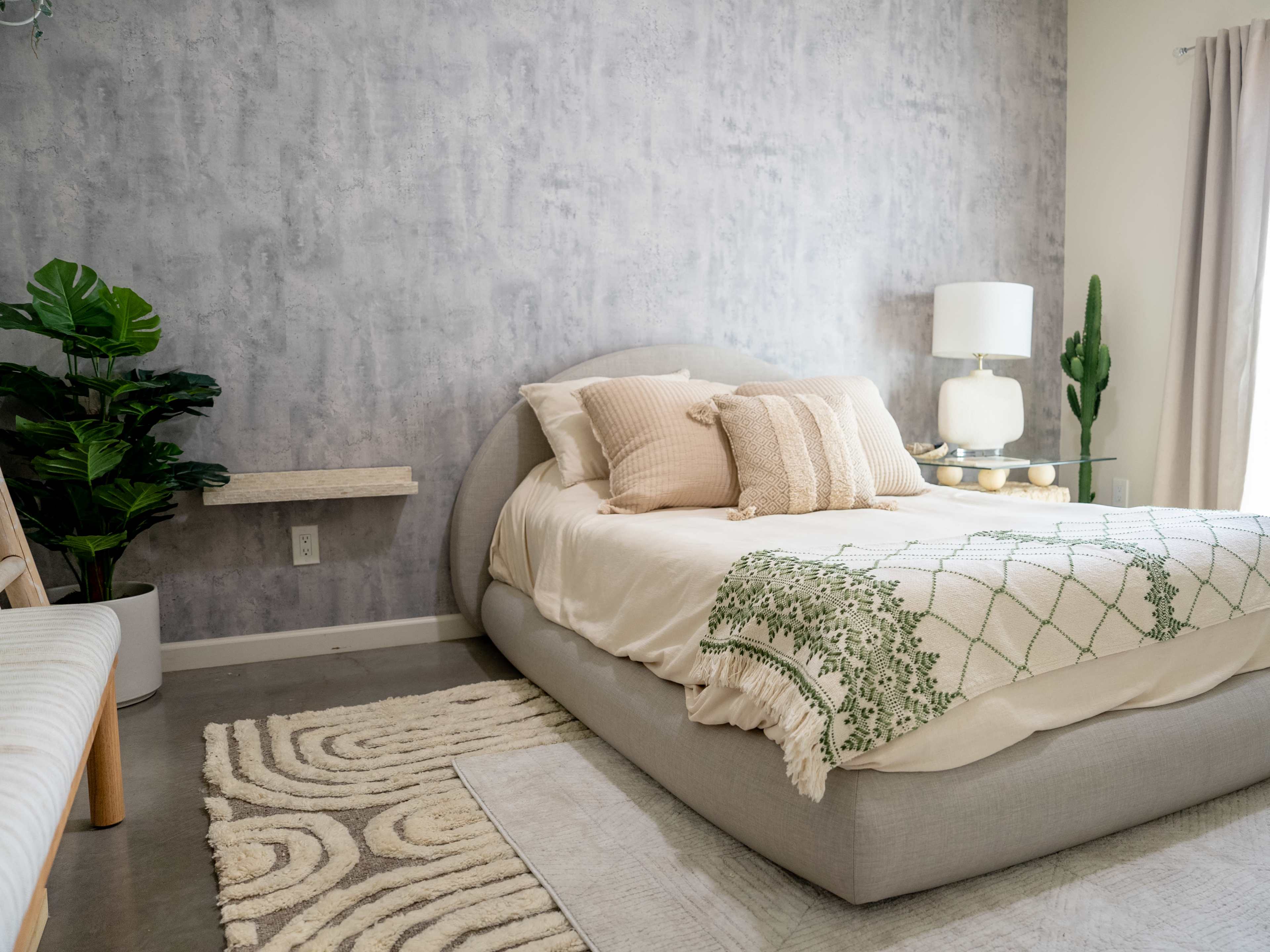 A modern bedroom featuring a curved bed with decorative pillows, a glass lamp on a side table, a small shelf, and green plants against a textured gray wall.