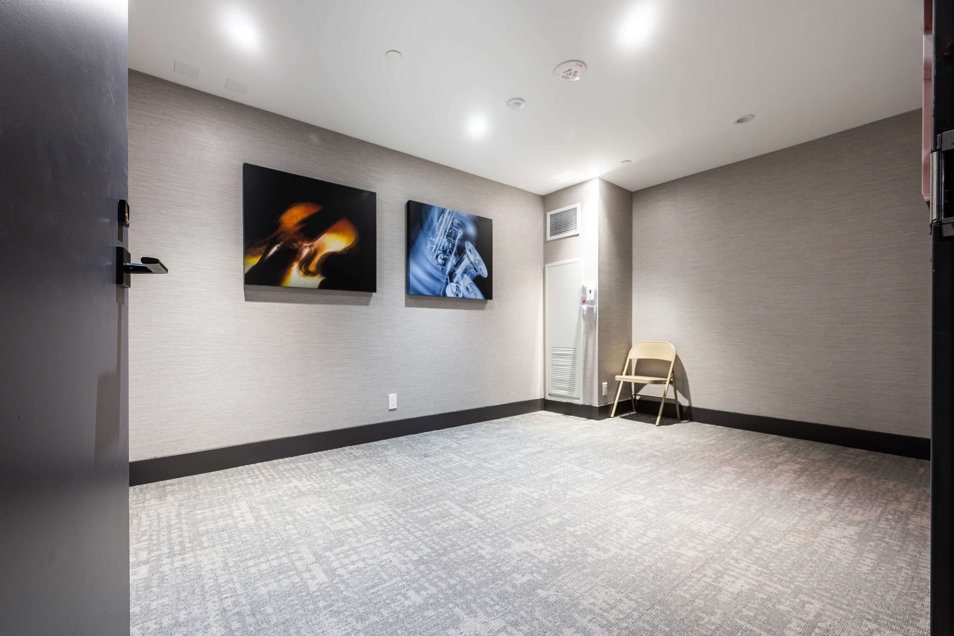 The image shows a small, empty room with two framed artworks on the wall and a single folding chair positioned against the wall.