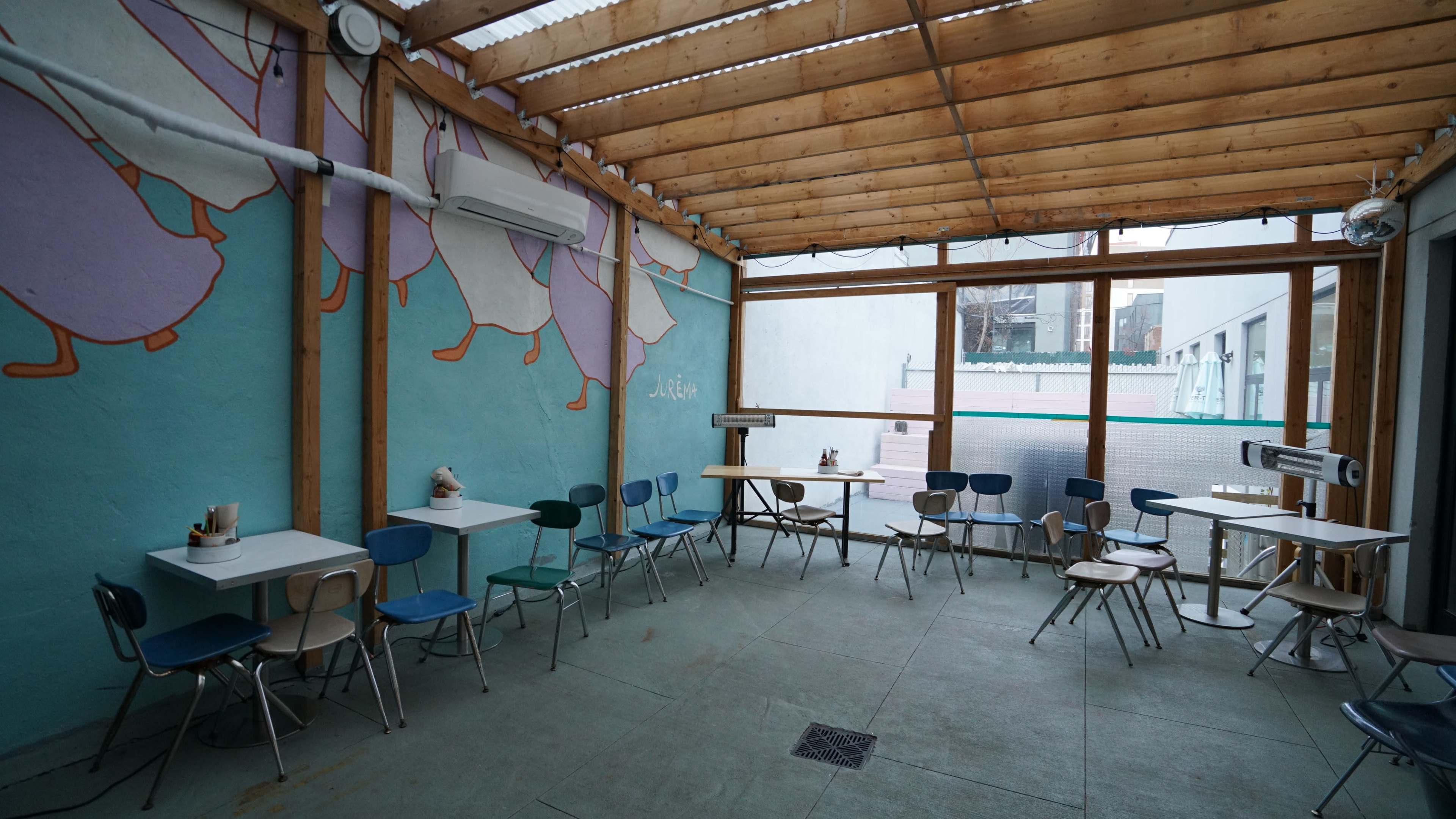 The image shows an indoor seating area with wooden beams and blue and beige chairs arranged around white tables against a colorful mural on the wall.