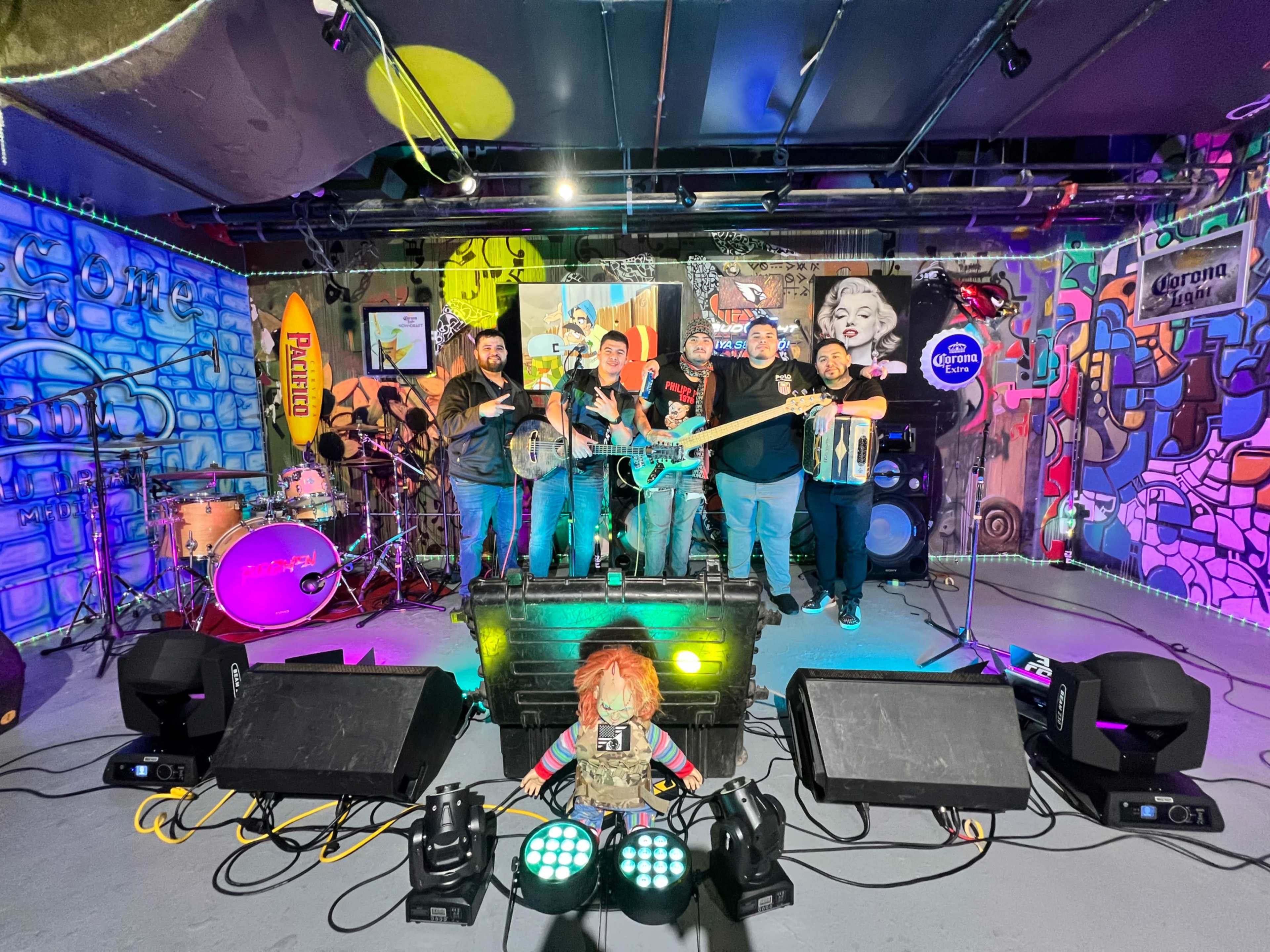 A group of five musicians poses on stage in front of a colorful, graffiti-covered backdrop with musical instruments and lighting equipment.