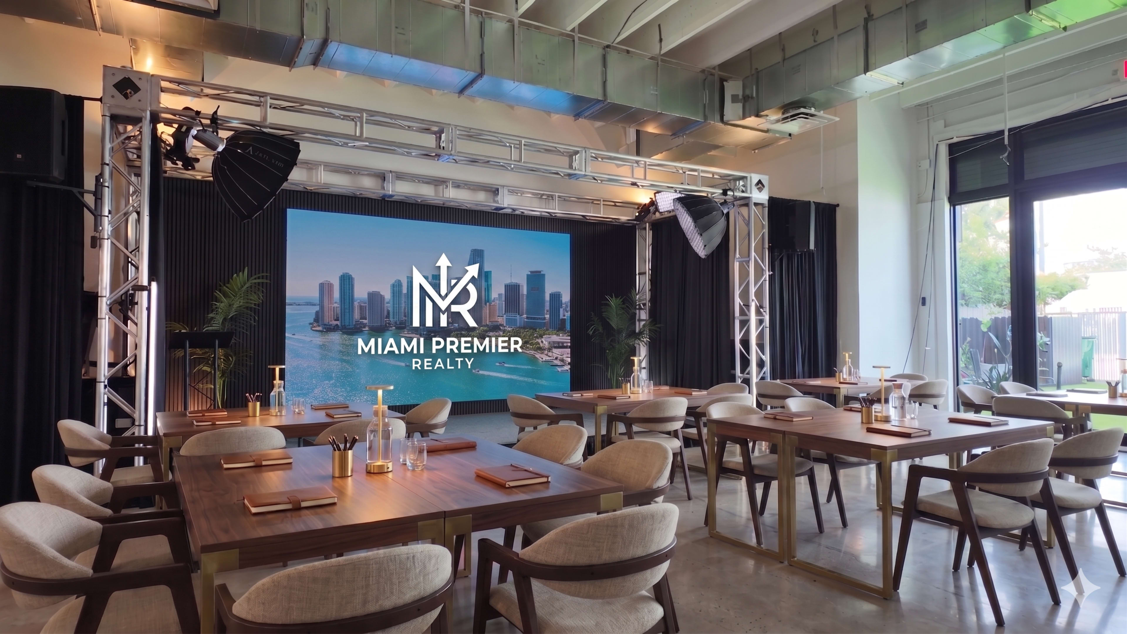 The image shows a modern event space set up for a presentation, featuring rows of wooden tables and a large screen displaying the "Miami Premier Realty" logo, with a view of Miami's skyline in the background.