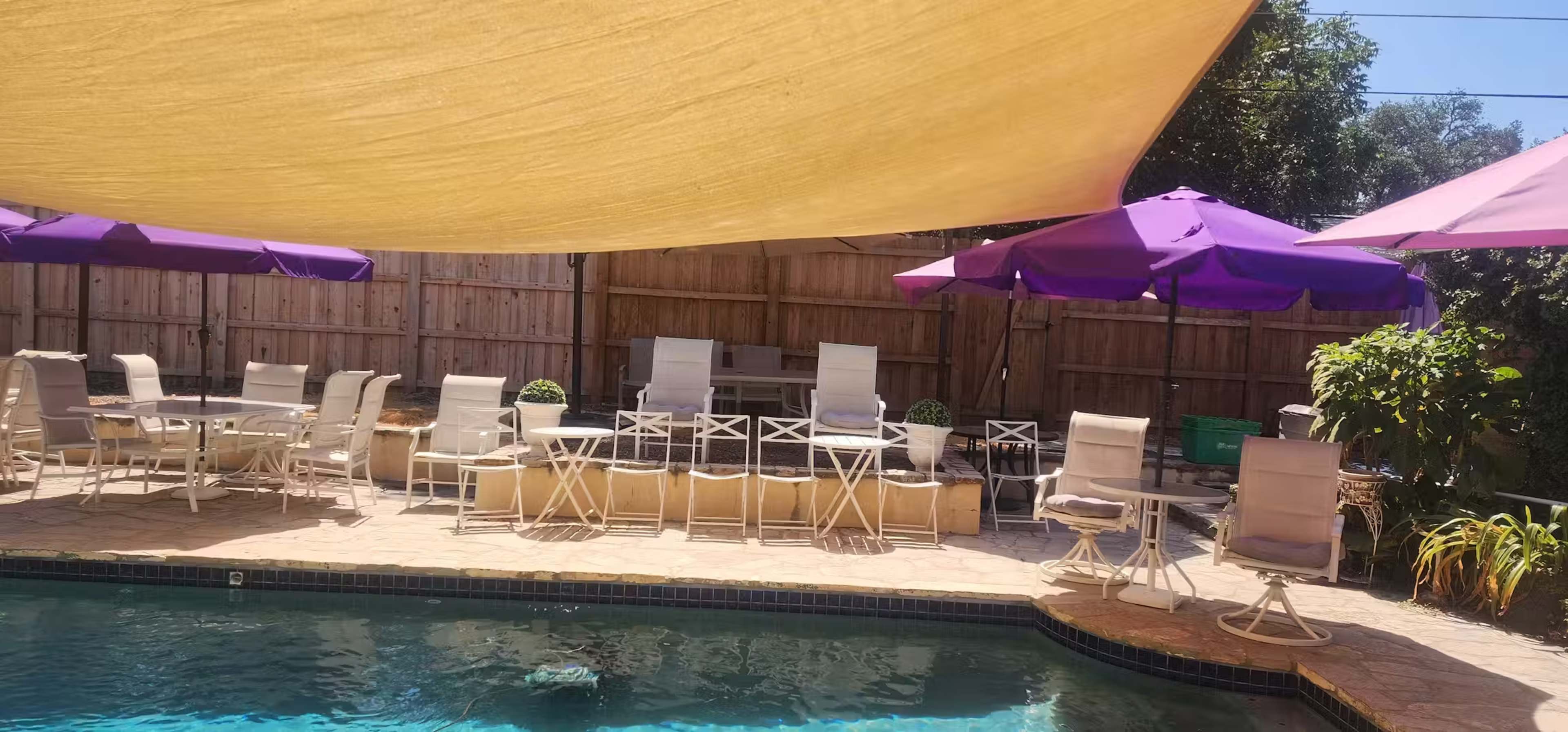 The scene shows a backyard pool area with a shaded seating area featuring white chairs and purple umbrellas.