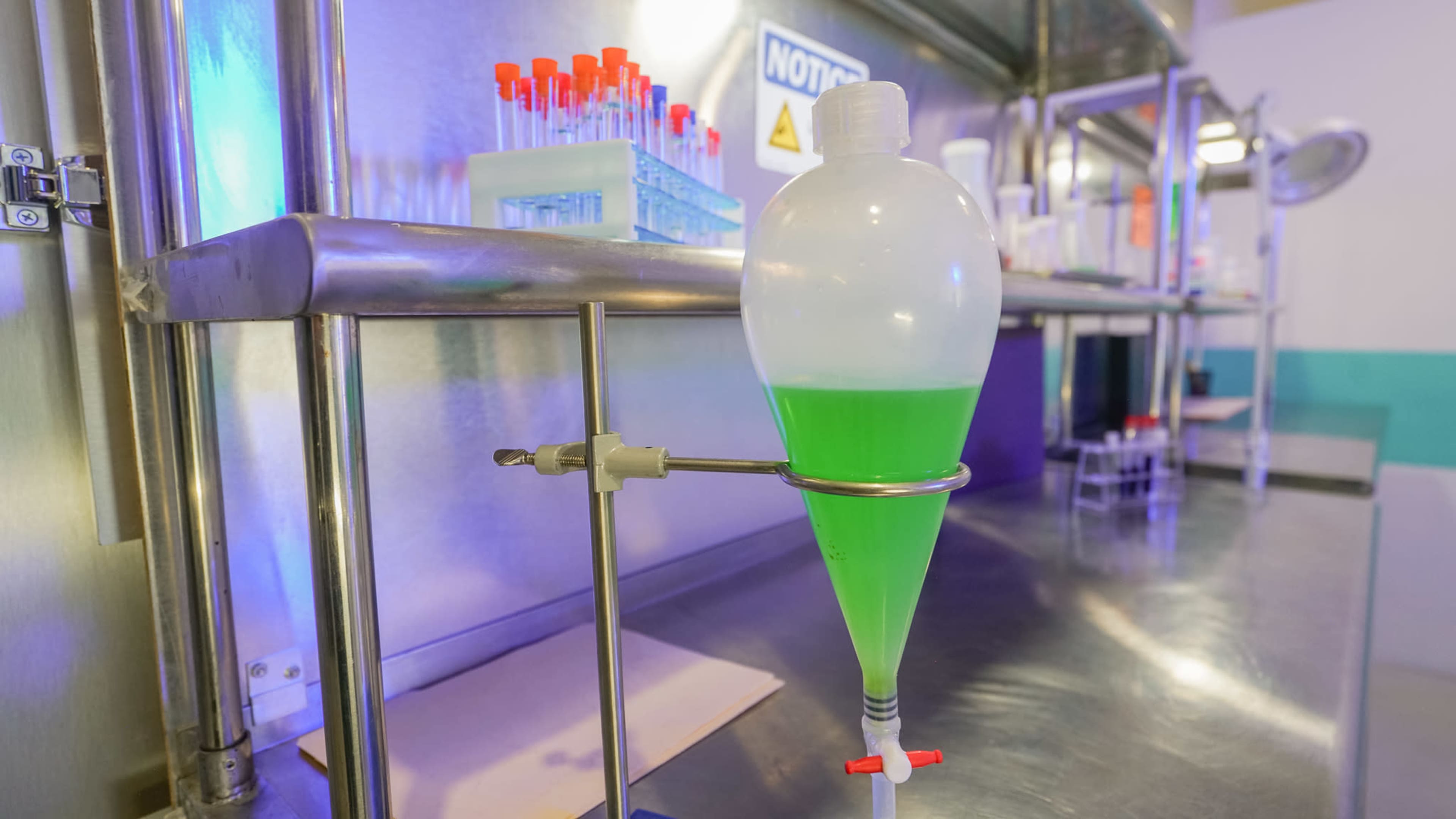 A green liquid is held in a glass funnel attached to a metal stand in a laboratory setting.