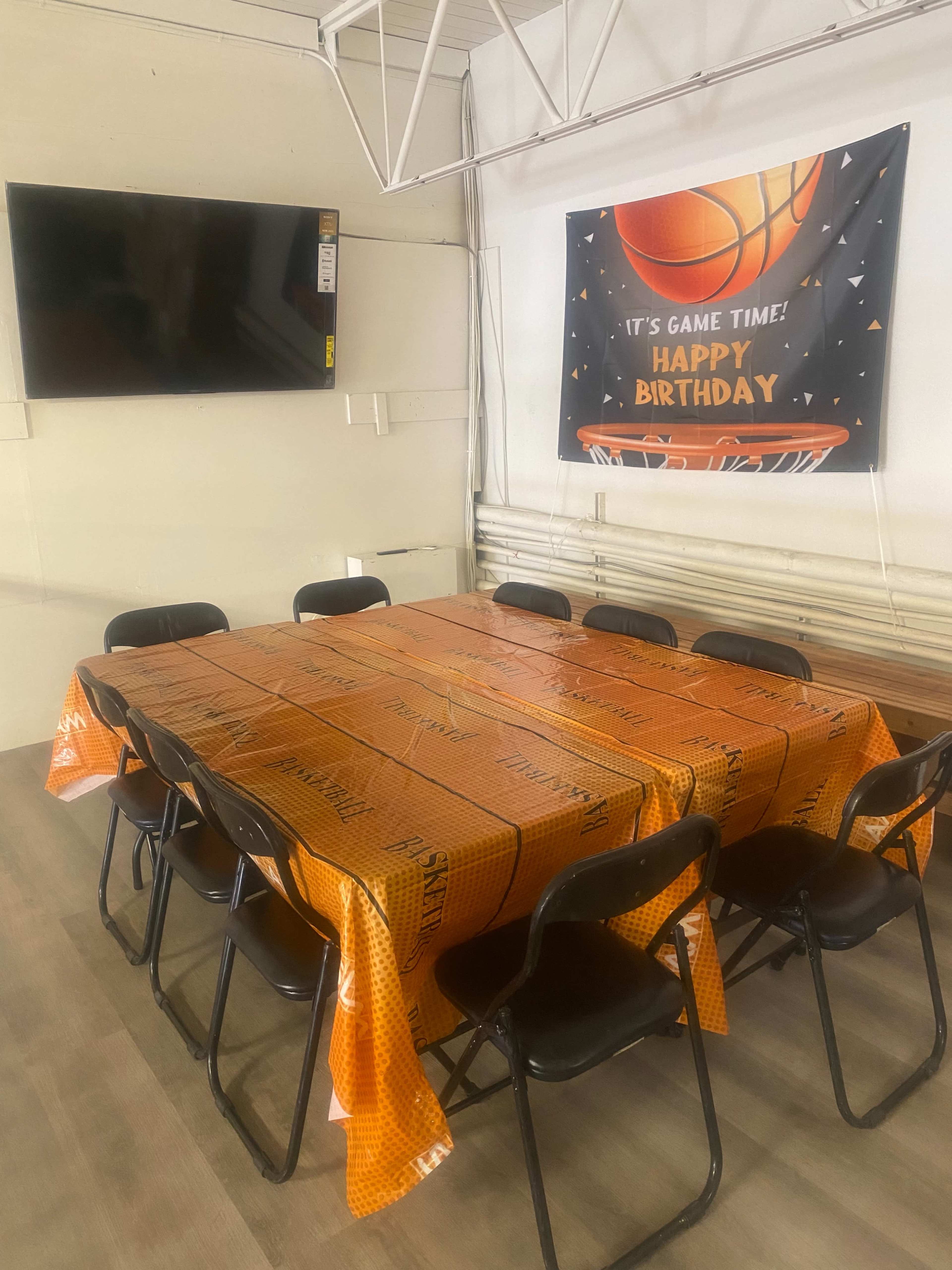 A rectangular table covered with an orange basketball-themed tablecloth is surrounded by black chairs in a decorated room featuring a "Happy Birthday" banner and a television on the wall.