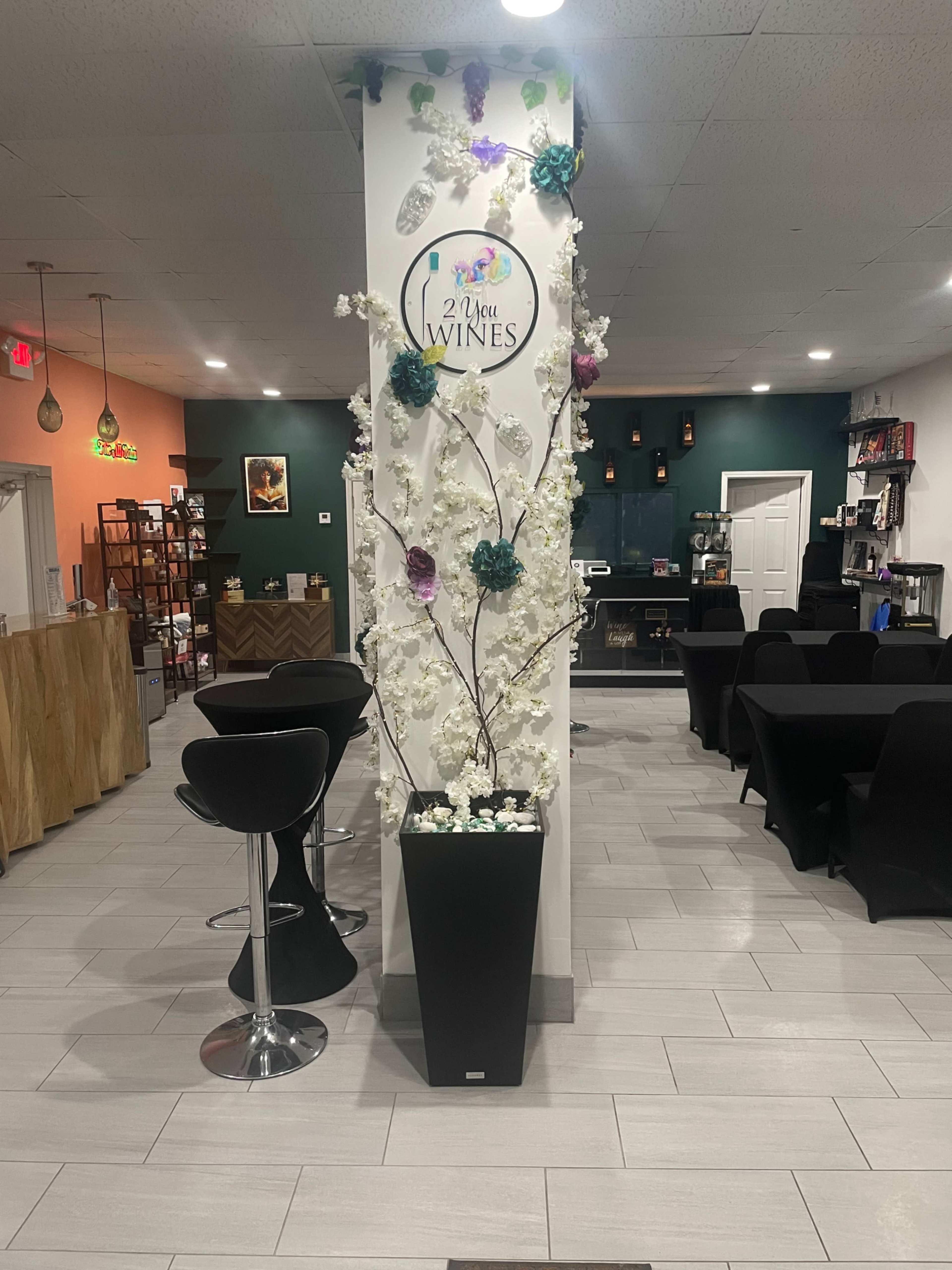 The image shows a modern interior of a wine bar, featuring a decorative display with artificial flowers and the sign "2 Wine Wines" at the center, surrounded by seating areas and a bar counter.