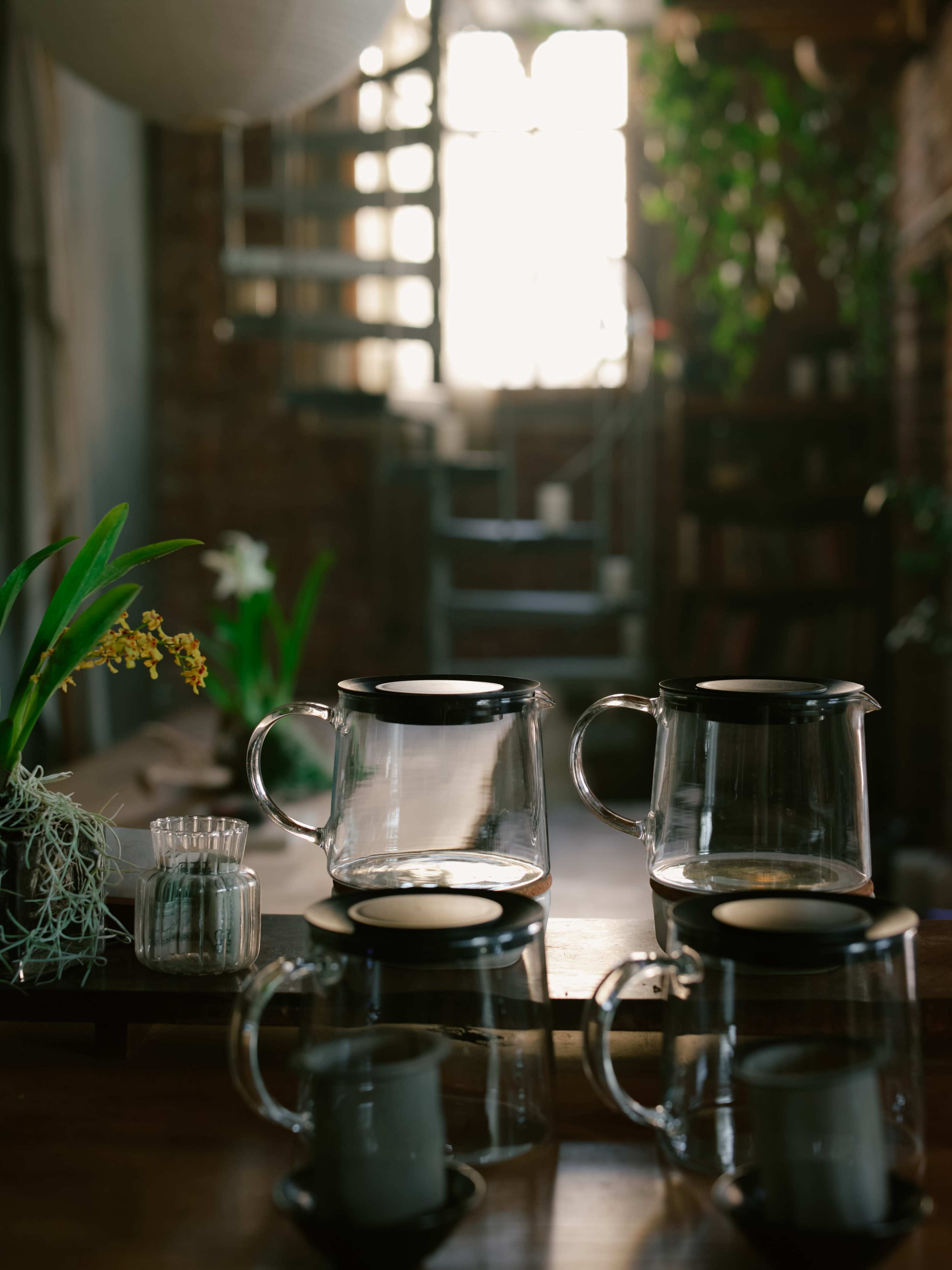 The image shows a collection of glass teapots on a wooden countertop, with a staircase and plants visible in the background.