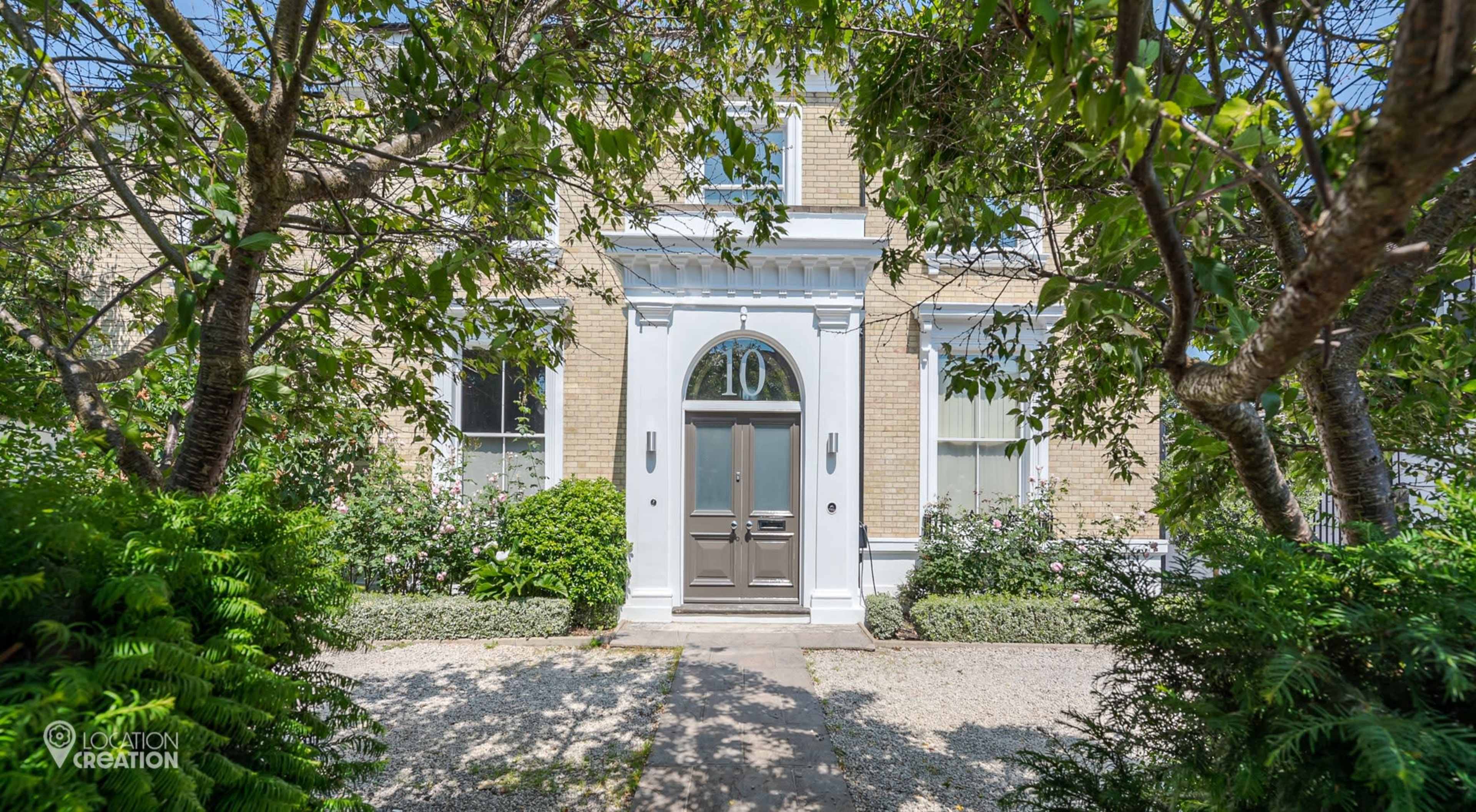 The image shows a house with a front entrance featuring a large door framed by a white architectural design, surrounded by lush greenery and a gravel pathway.