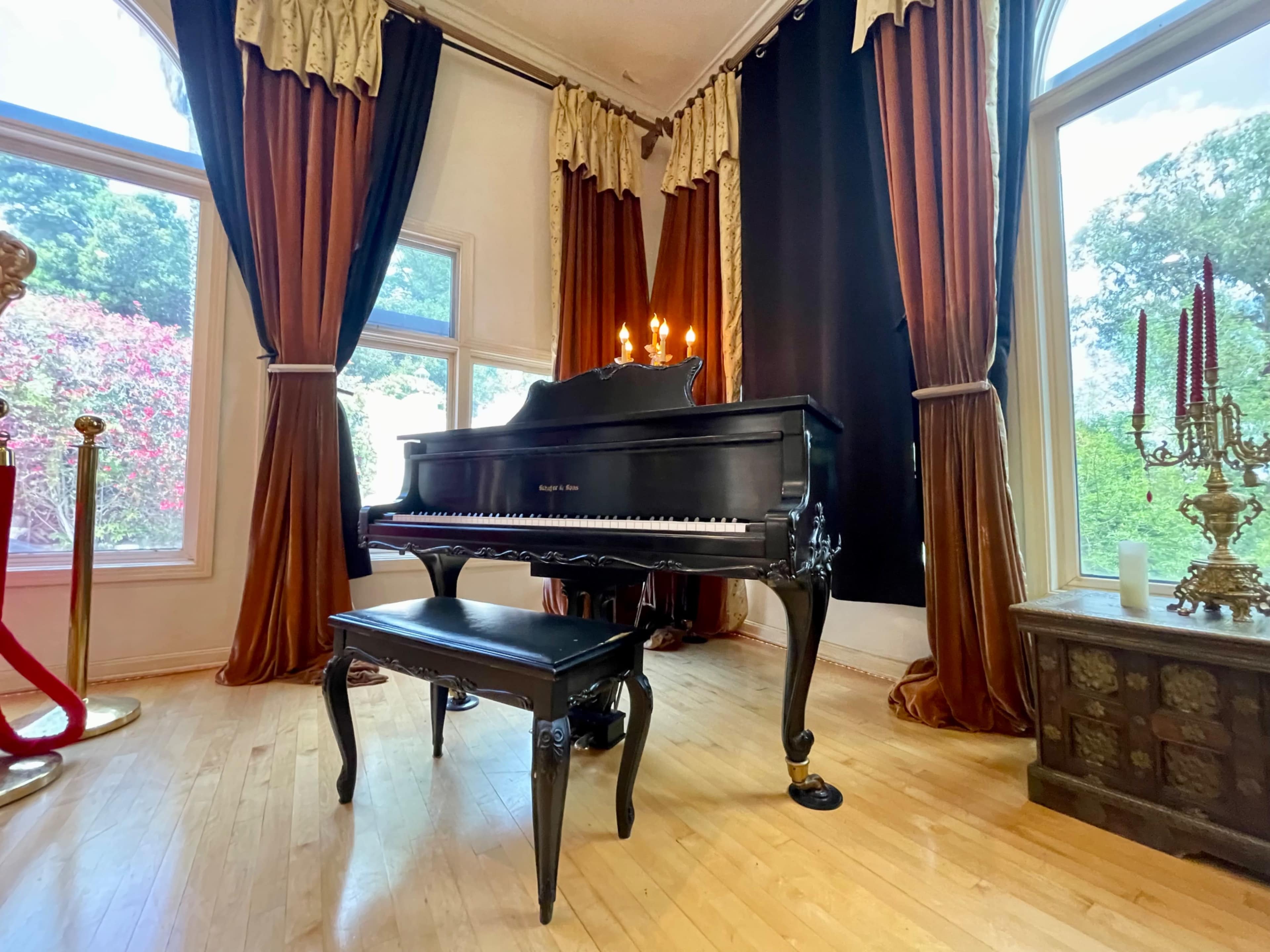 A black grand piano with a matching bench is positioned in a well-lit room featuring large windows and elegant drapery.