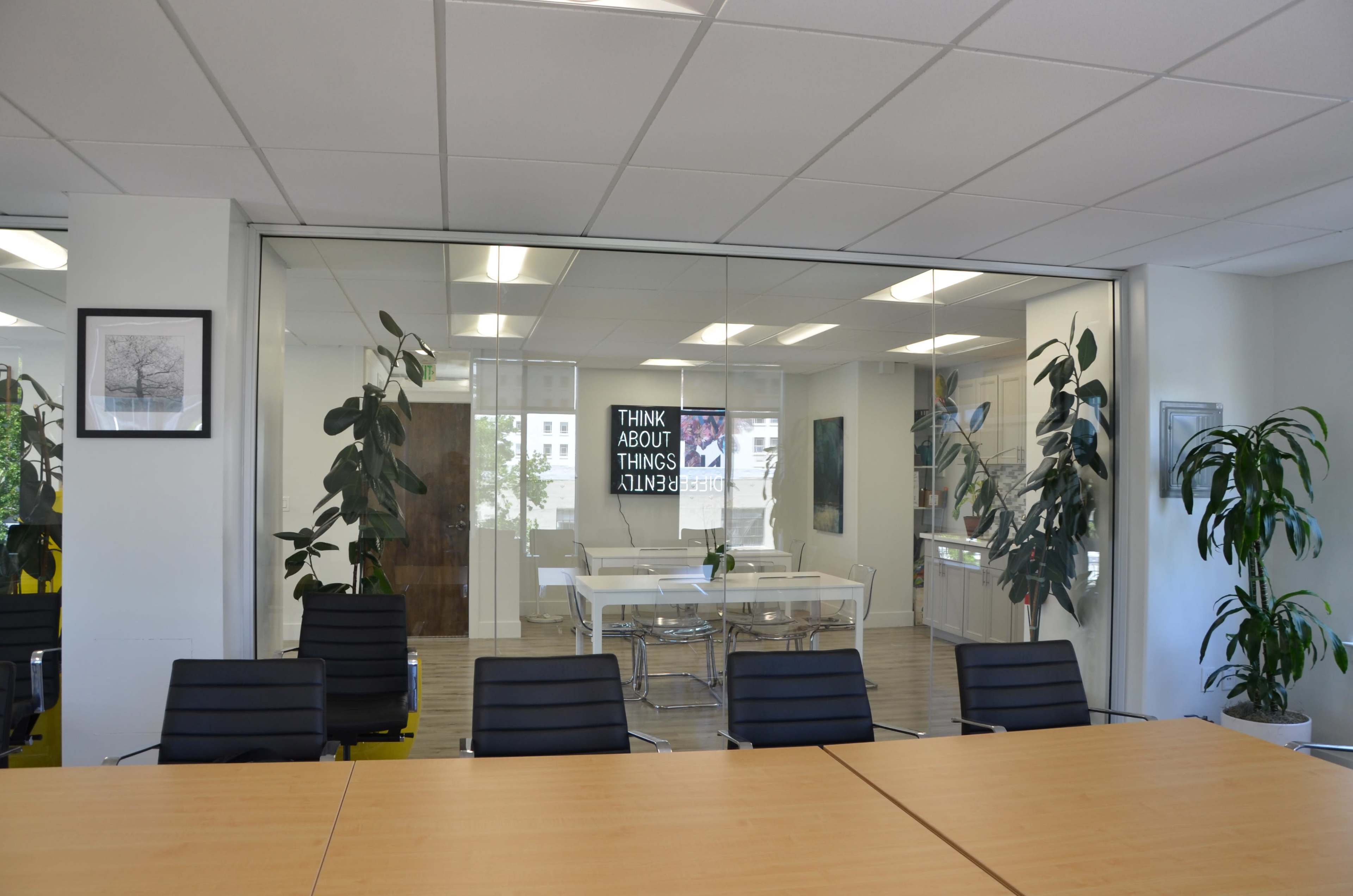 The image shows a modern office area with a glass-walled conference room featuring a large table, black chairs, and greenery, alongside motivational text on the wall.