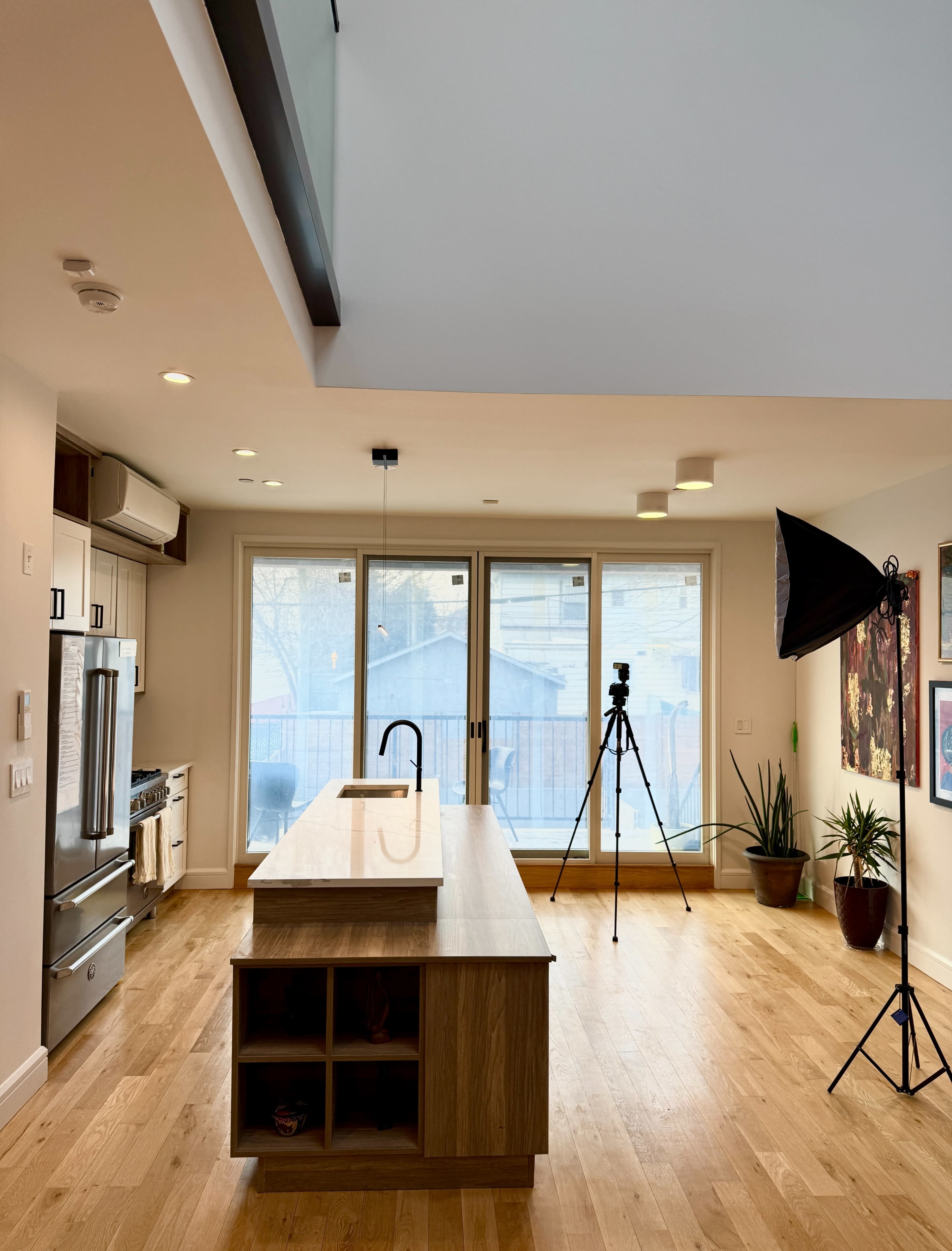 The image shows a modern kitchen with a wooden island, large windows, and a tripod set up for photography.
