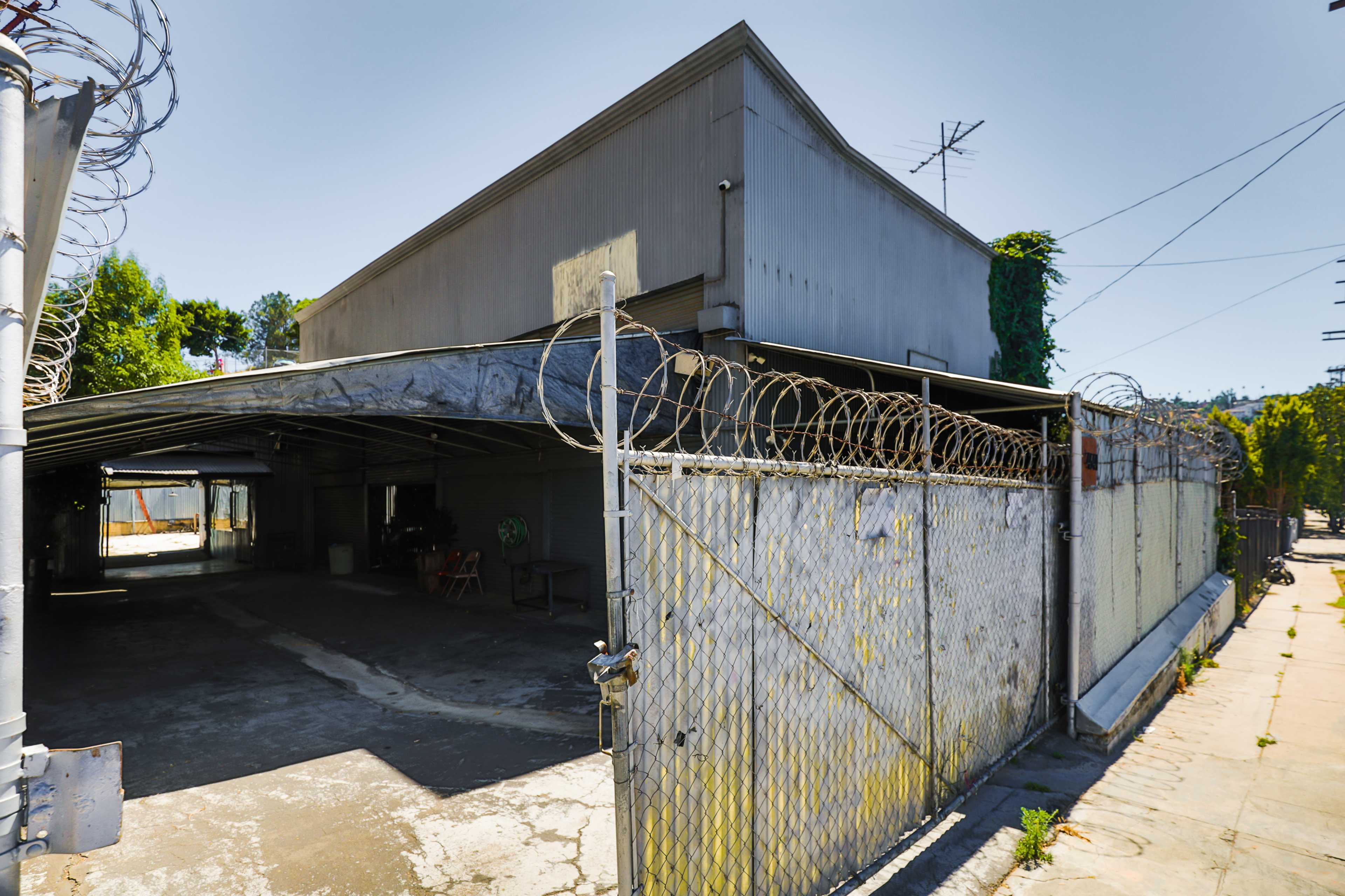 A metal fence topped with barbed wire surrounds a gray building with a flat roof and an empty driveway, set against a clear blue sky.