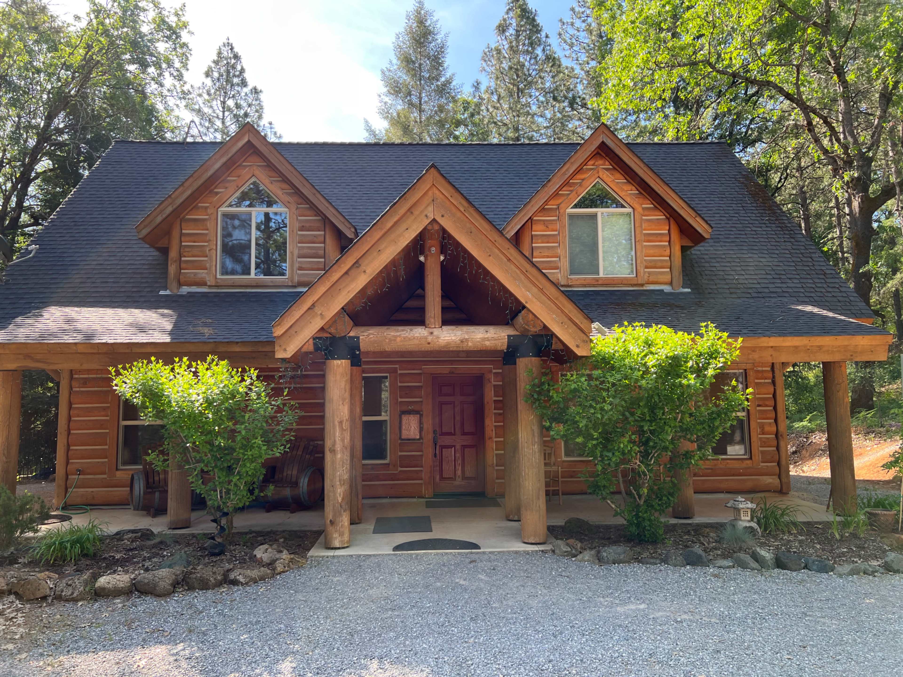 The image shows a large log cabin with a peaked roof, surrounded by trees and a gravel driveway.