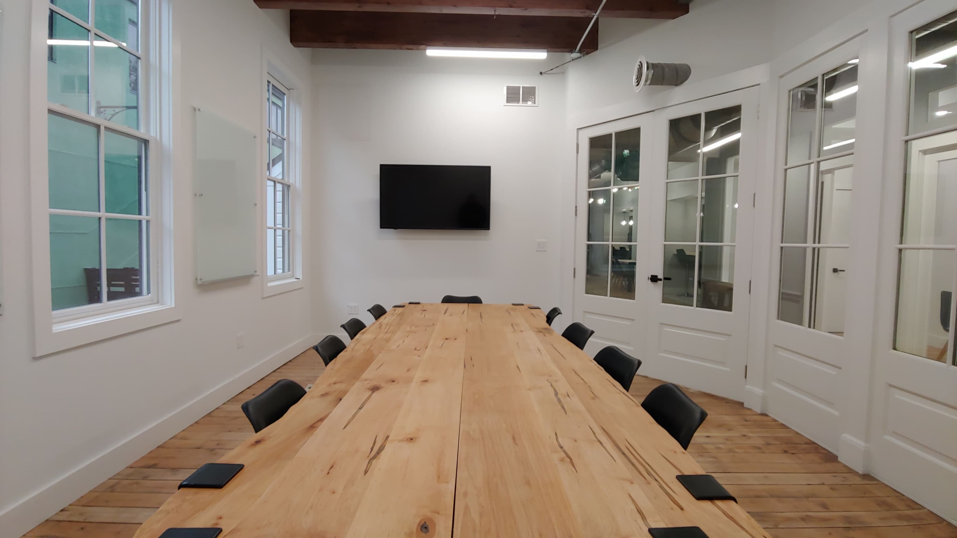 A long wooden table is set up in a brightly lit conference room, with chairs arranged around it and a large screen mounted on the wall.