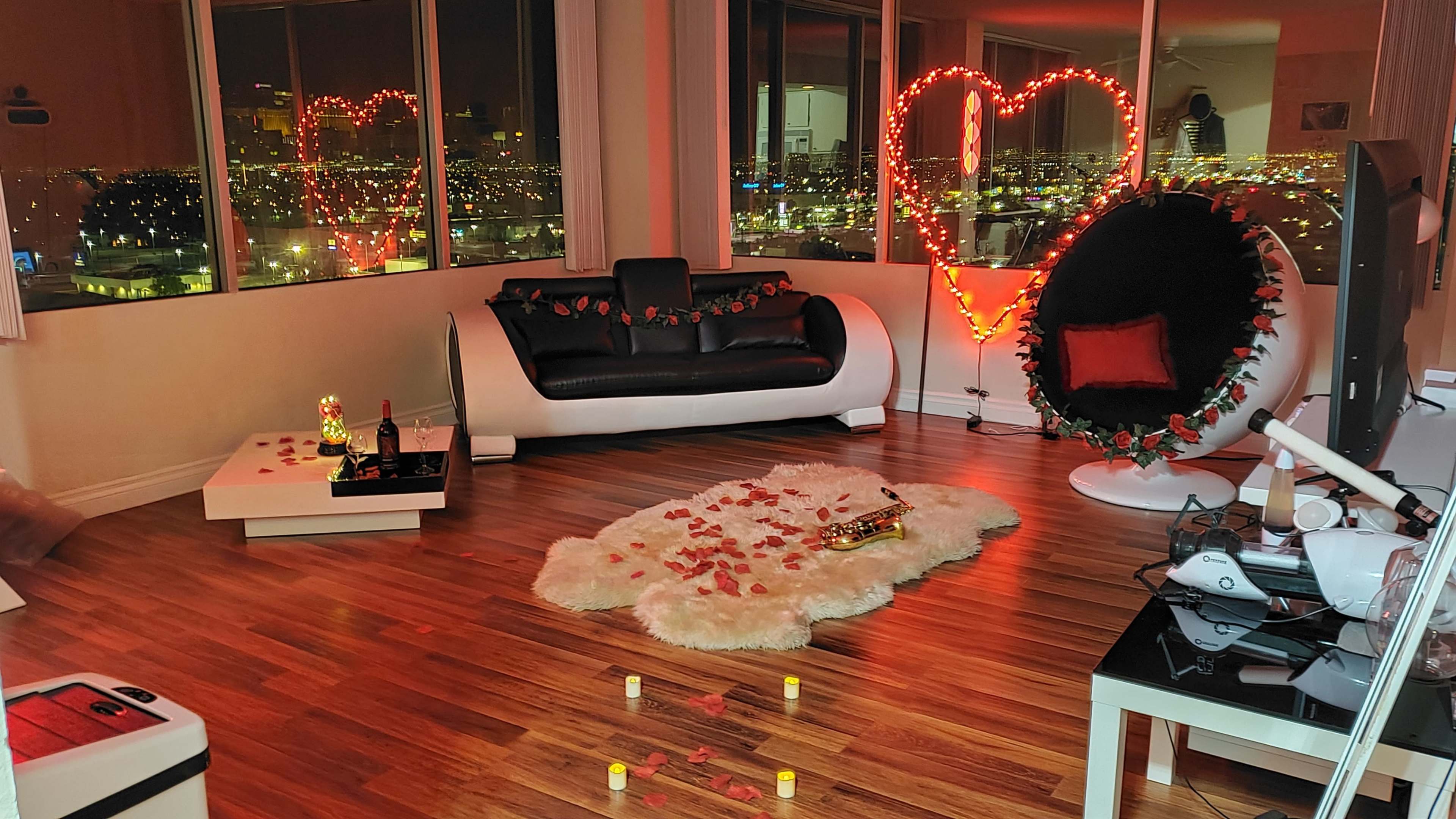 The room features a modern seating area with a black and white couch, a round chair, a heart-shaped light display, and decorative elements like rose petals and candles on the floor, with city lights visible through large windows.