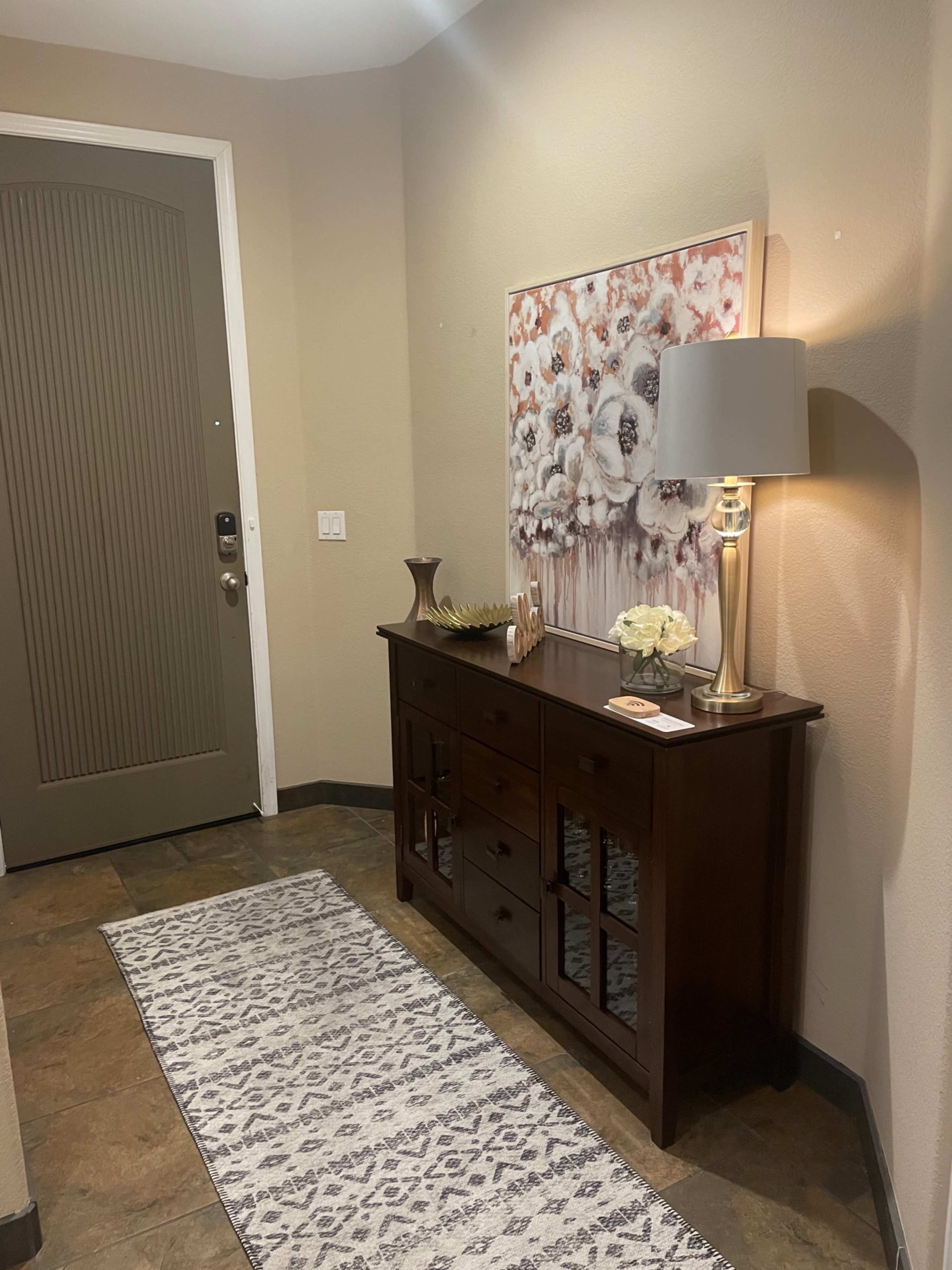 A small entryway features a dark wooden console table with decorative items and a table lamp against a light-colored wall, with a floral artwork hanging above and a green front door visible.