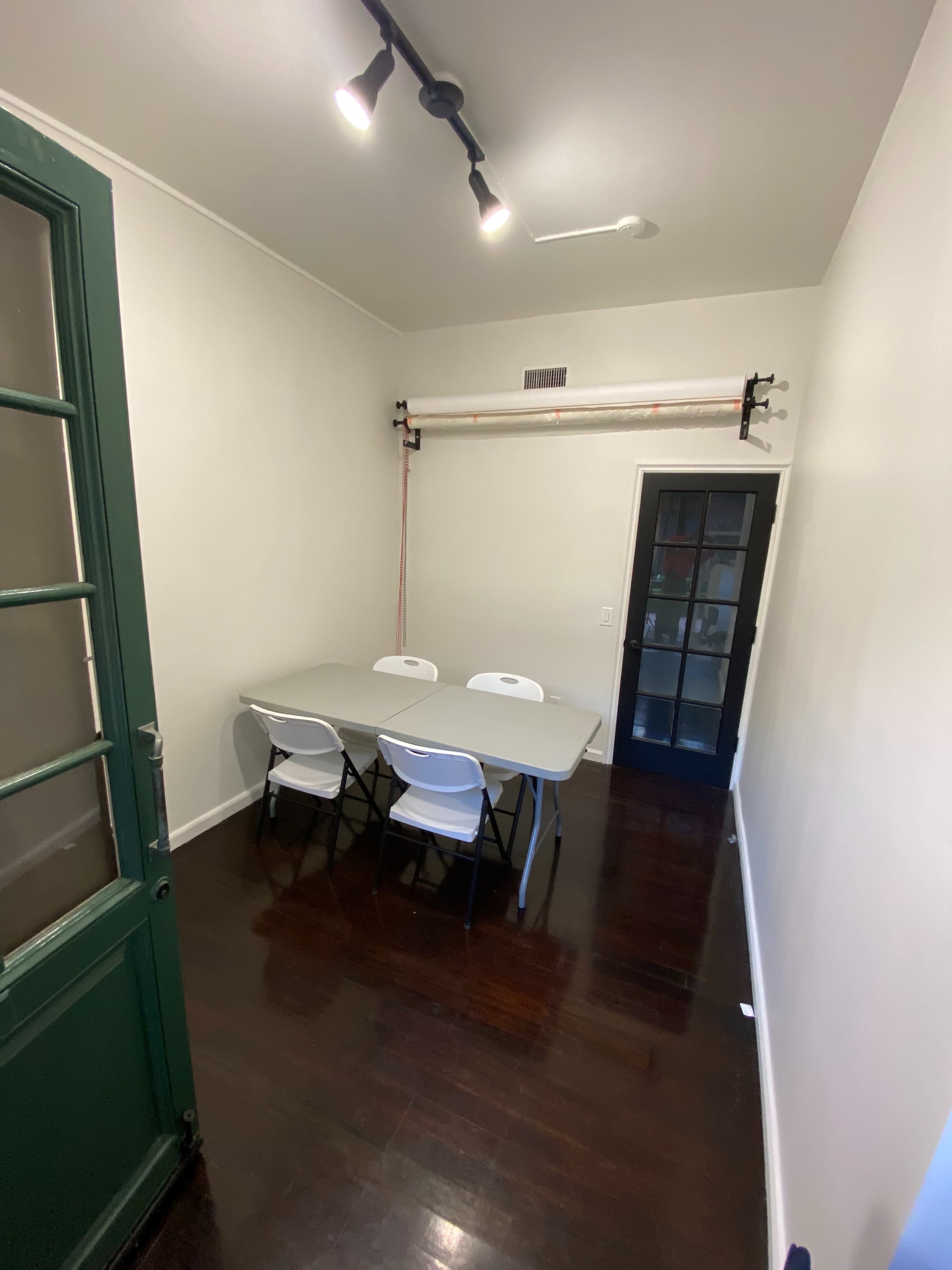 The image shows a small room with a green door, a table surrounded by four white chairs, and a black-framed glass door leading to another space.